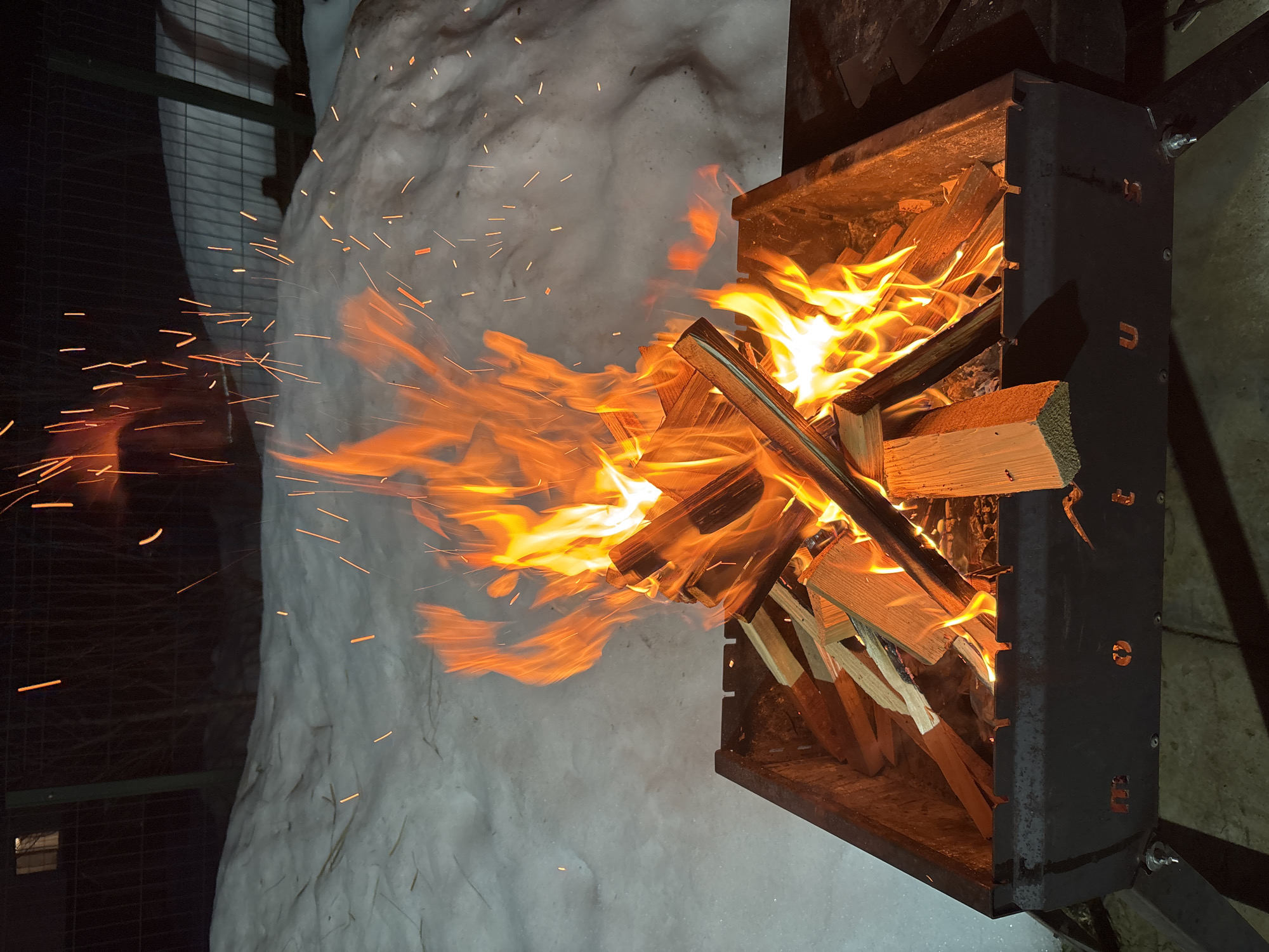 This image depicts a nighttime outdoor scene in Русский Вожой, Russia. A rectangular black metal brazier (mangal) is centrally positioned, containing an active wood fire. Bright orange and yellow flames rise from several pieces of split firewood within the brazier, emitting numerous glowing sparks that ascend into the dark atmosphere. The ground immediately surrounding the brazier is covered in an uneven layer of white snow. In the dark background, a wire or mesh fence is visible, and beyond it, a faint building with one illuminated window can be observed. No individuals are present in the scene.