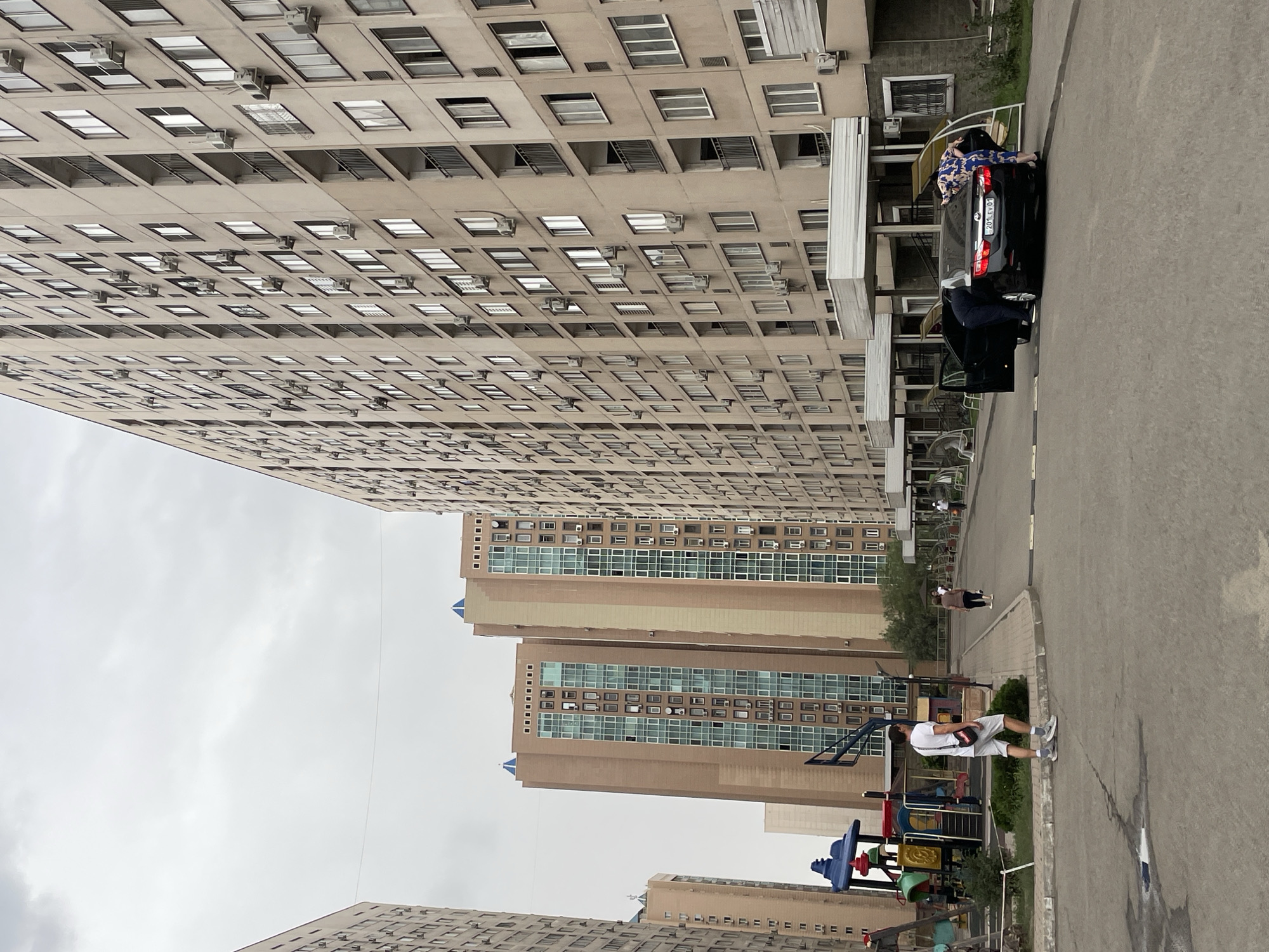 A wide shot captures a street scene in Almaty, Kazakhstan, with towering apartment buildings dominating the background. On the left, a colorful playground features a slide and swings. A man in a white t-shirt and shorts stands in the foreground, facing away from the camera, with a black crossbody bag. Further down the street, a black sedan is parked with its doors open, and two individuals are interacting with the vehicle. One person, wearing a dark suit, is leaning into the car, while another in a patterned dress stands near the trunk. The sky is overcast, casting a diffused light on the urban landscape, which includes a mix of concrete and brick architecture.