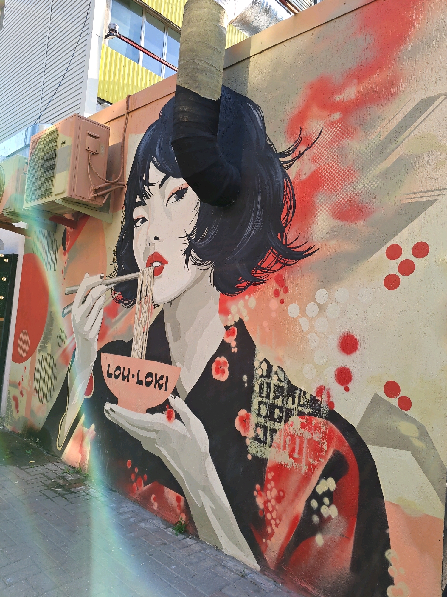 The image shows a mural on a wall in Rostov-on-Don, Russia, depicting an Asian woman with dark hair eating noodles with chopsticks. The woman is wearing a black garment with red floral accents and has bright red lipstick. A pink bowl with the text "LOU LOKI" is held by her hands. The mural is done in a graphic, stylized manner with bold lines and splashes of red and white paint. Behind the woman, there are abstract red and white circular shapes and a grid pattern. To the left of the mural, an air conditioning unit is mounted on the wall. The mural is located on an exterior wall, and the ground in front of it is paved with grey bricks. A rainbow-like lens flare effect is visible in the lower left quadrant of the image.