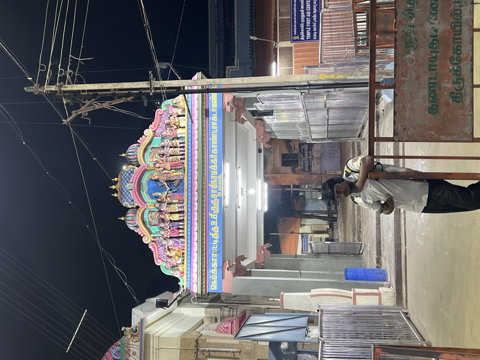The image, taken at night in Palani, India, depicts the brightly illuminated entrance of a Hindu temple. On the right side of the frame, a man in a light-colored uniform shirt and dark trousers leans against a metal barrier, looking towards the camera. He appears to be a guard or temple staff member.
The prominent feature on the left is a colorful, multi-tiered temple gopuram (tower), intricately decorated with numerous sculptures of deities and mythological figures. The vibrant colors of the gopuram are highlighted by artificial lighting against a dark night sky. Below the gopuram is an ornate archway, serving as the temple entrance, also adorned with sculptures and an inscription.
Overhead, a complex network of electrical wires crisscrosses the sky, connected to a utility pole, indicating an urban or semi-urban setting. Several signs with text are visible: one explicitly states "TEMPLE FIRST AID CENTER" with its Tamil equivalent "திருக்கோயில் முதலுதவி மையம்". Another sign identifies the temple as "தண்டாயுதபாணி திருக்கோயில்" (Dhandayuthapani Temple). An inscription on the temple archway reads "நெய்க்காரபட்டிதிரு.உ.சி.குருசாமிநாயக்கர்காண்ட்ராக்டர்மண் உபயம்," likely a donor's name or sponsorship detail. A blue plastic barrel is on the ground in the midground. The scene portrays a calm, active temple environment after dark.