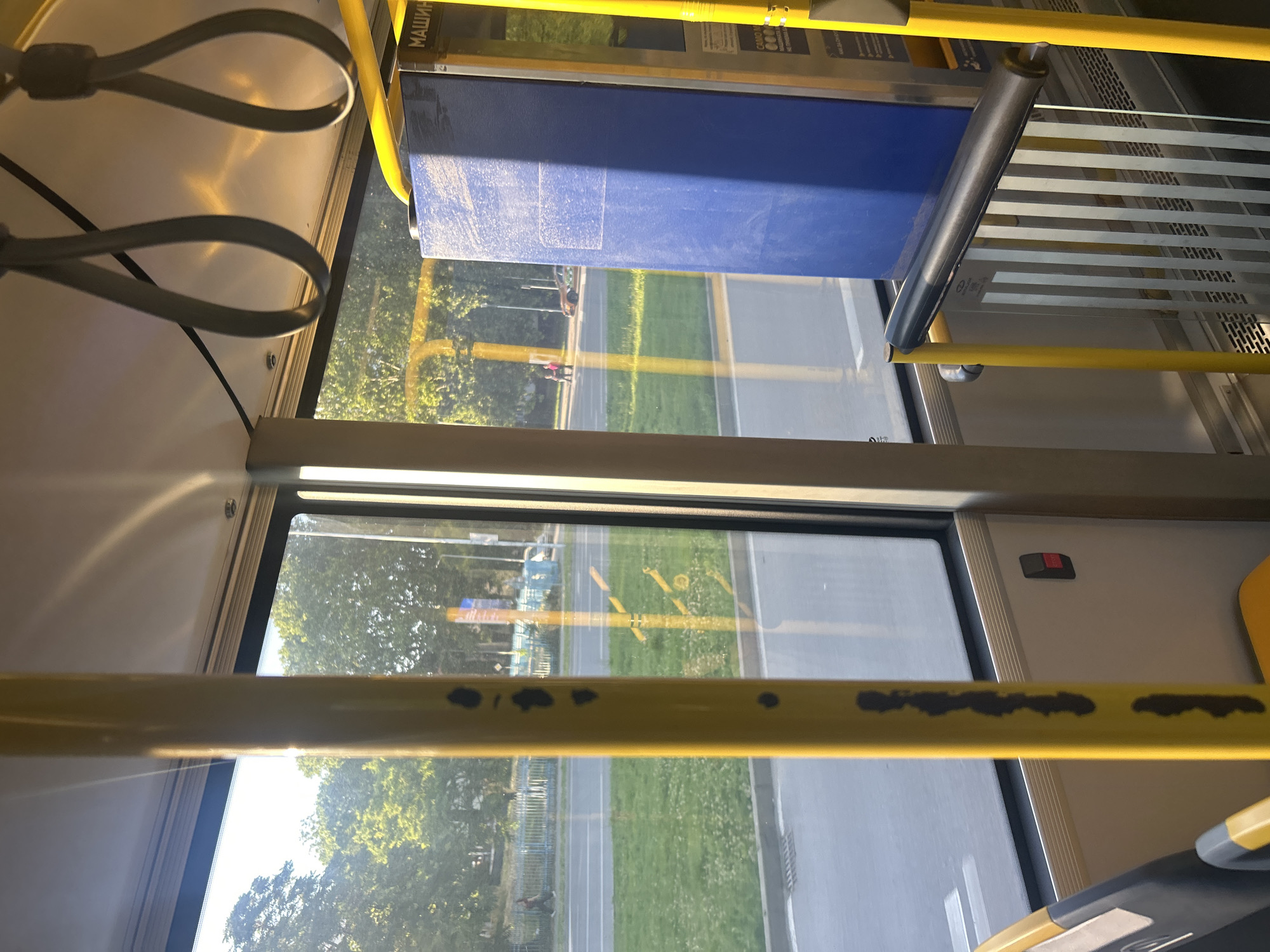 The image shows the interior of a public transportation vehicle, likely a bus or tram. Yellow poles and handrails are prominent features, indicating a focus on passenger safety and mobility. Large windows offer a view of a street scene with trees, grass, and a sidewalk. Sunlight streams in, creating bright reflections on the windows and interior surfaces. A large blue panel, possibly a ticket machine or information display, is visible on the right side of the image. The overall impression is one of a functional and utilitarian space, typical of public transport. There are no discernible people or specific actions occurring within the vehicle or immediately outside. The urban environment appears green and well-maintained.