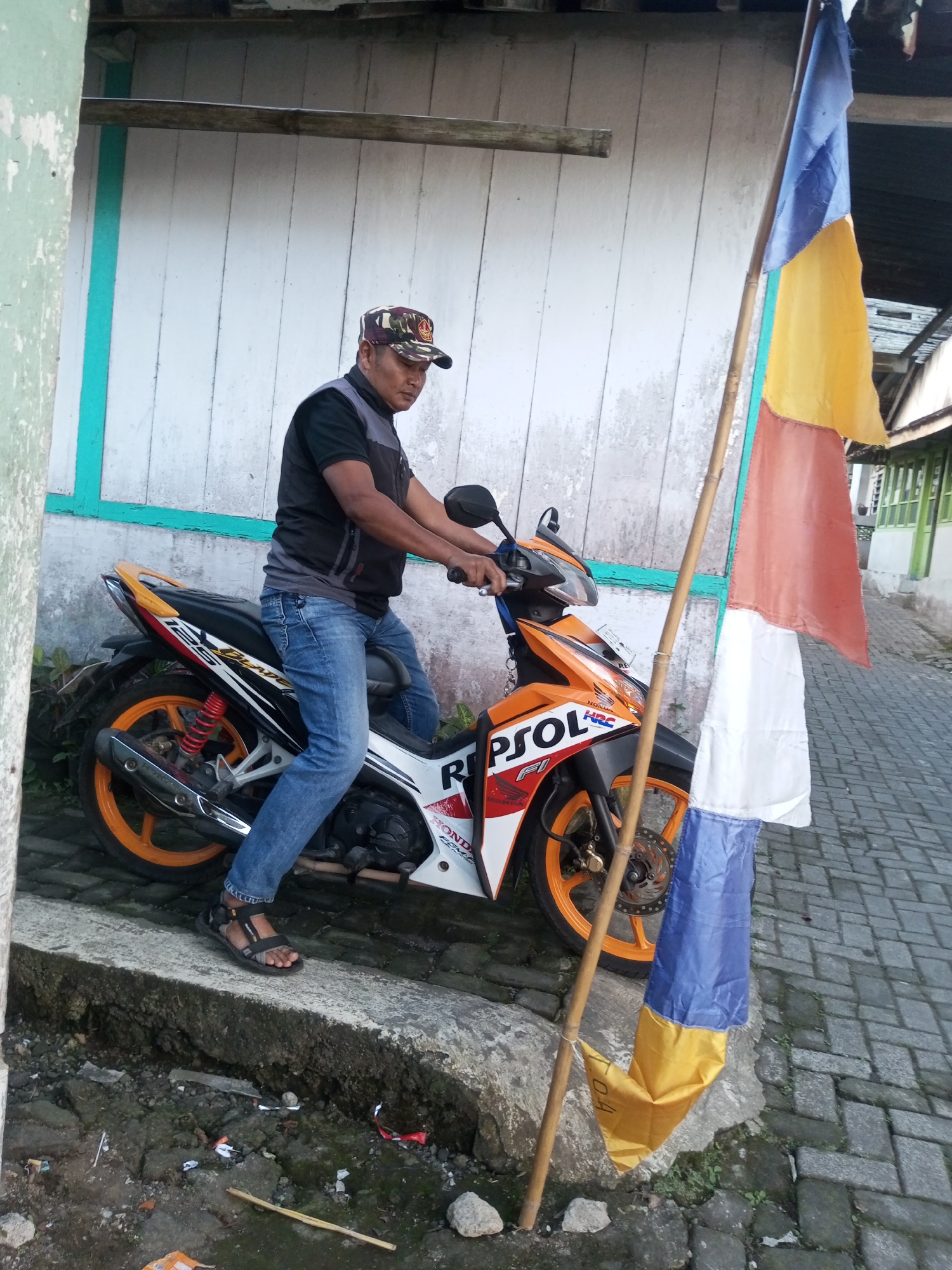 A man sits on an orange and white motorcycle, facing right, in the city of Andongsili, Indonesia. He is wearing a camouflage baseball cap, a black and gray vest over a black shirt, and blue jeans. His feet are on the ground, with one foot resting on a curb. A multi-colored flag, with blue, yellow, orange, and white sections, is planted in the ground to the right of the motorcycle, held by a bamboo pole. The motorcycle has a "REPSOL" logo on its side. The background consists of a light-colored wooden wall and a paved area. The scene appears to be outdoors, with some greenery visible in the lower left corner.