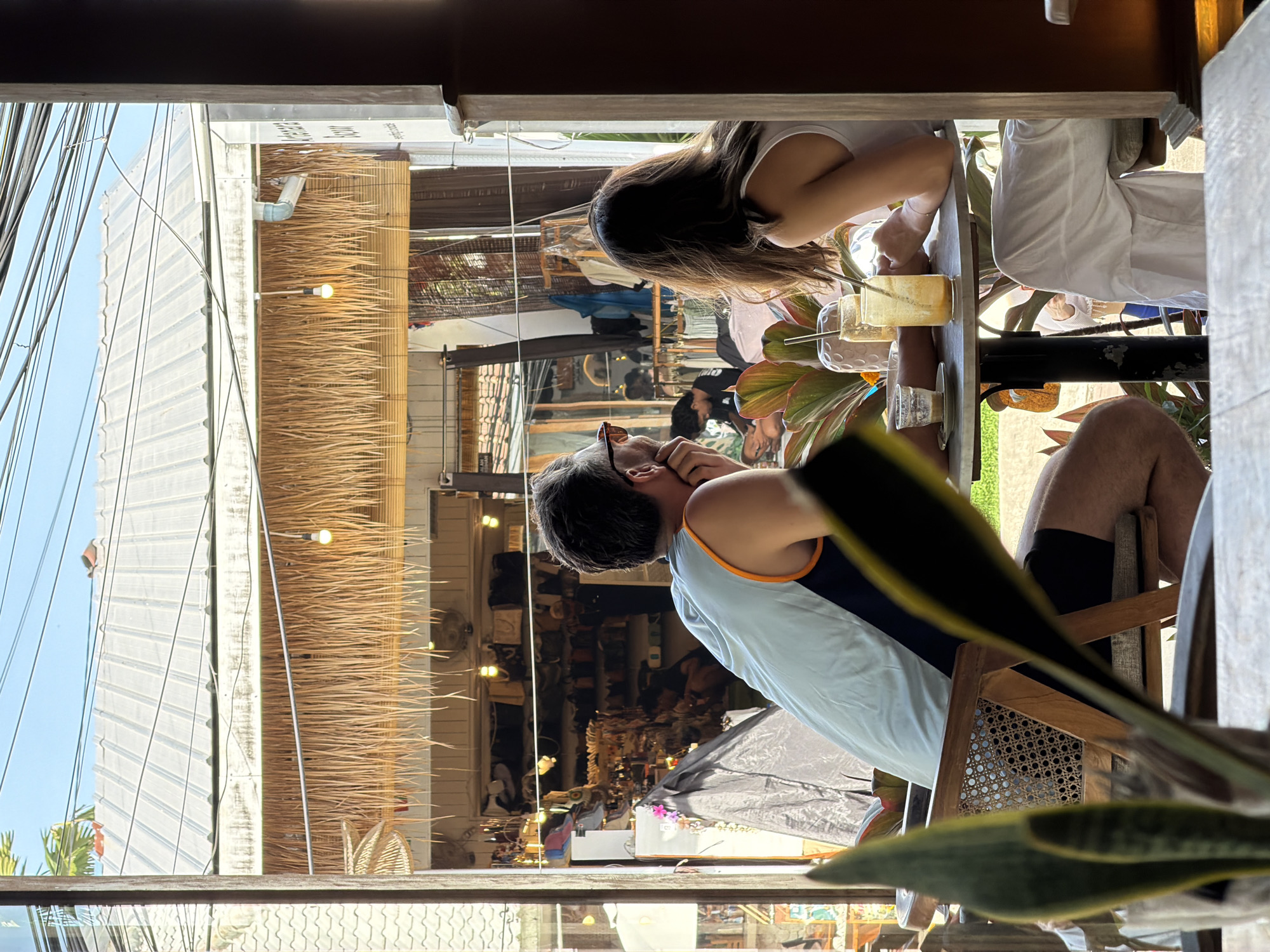 A partially occluded interior shot reveals a man and a woman seated at a table, observing a market street scene in Kuta Selatan, Indonesia. The man, wearing a light blue tank top with orange trim, has dark hair and is looking through sunglasses towards the market. The woman has long dark hair and wears a white tank top. On the table between them are two drinks, possibly containing ice, and a small vase with pink flowers. The market street is lined with stalls under thatched roofs. A variety of goods appear to be displayed. Power lines run across the upper left of the view. A potted plant is in the foreground, partially obscuring the view.