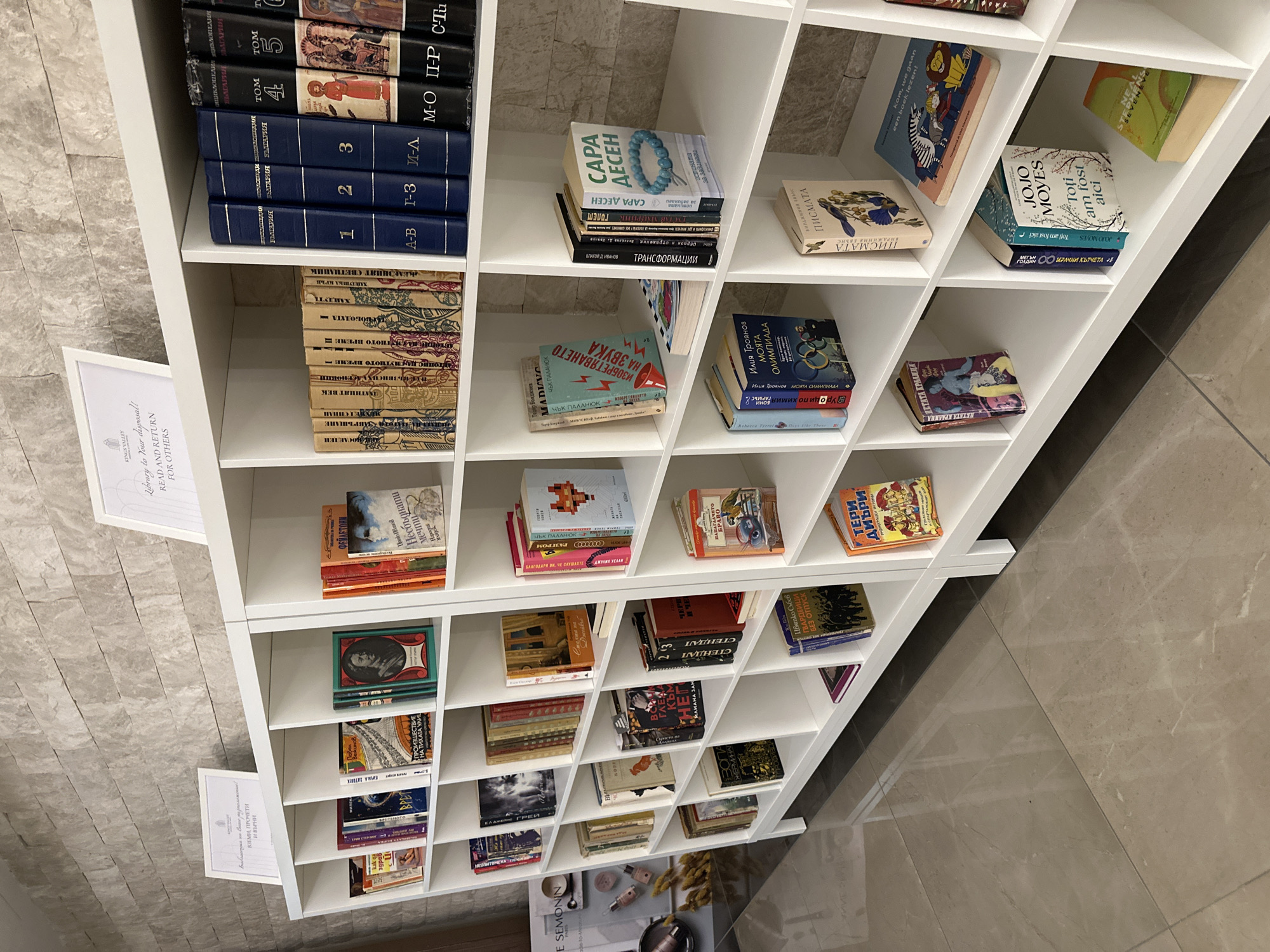 This image displays a brightly lit, white modular bookshelf filled with numerous books, suggesting a community library or book-sharing point in Kazanlak, Bulgaria. The books are arranged in a mix of horizontal stacks and vertical displays within the square compartments of the shelving unit.
The background features a light-colored, textured stone or tile wall, while the floor below is a smooth, light-toned tile with visible seams.
Two framed signs are visible on the left side of the shelving. The lower sign, clearly legible, states in English: "KINGS VALLEY Library to Your disposal! READ AND RETURN FOR OTHERS." The upper sign, also in a white frame, has Cyrillic text, prominently featuring "ВЗЕМИ, ПРОЧЕТИ И ВЪРНИ," which translates to "Take, Read, and Return," reinforcing the purpose of the book collection.
The books are predominantly in Bulgarian, indicated by the Cyrillic script. Notable visible titles and authors include works by Sarah Dessen ("САРА ДЕСЕН"), Jojo Moyes ("JOJO MOYES" with "Toți am fost aici" likely Romanian, meaning "We were all here"), Chuck Palahniuk ("ЧЪК ПАЛАНЮК," with "ИЗОБРЕТЯВАНЕТО НА ЗВУКА" - *The Invention of Sound*), Terry Deary ("ТЕРРИ ДИЪРИ"), Stendhal ("СТЕНДАЛ" with "ЧЕРВЕНО И ЧЕРНО" - *The Red and the Black*), and Iliya Troyanov ("Илия Троянов МОЯТА ОЛИМПИАДА" - *My Olympiad*). A multi-volume encyclopedia set titled "БЪЛГАРИЯ" (Bulgaria), numbered 1 through 5, is also visible, arranged by alphabetical ranges.