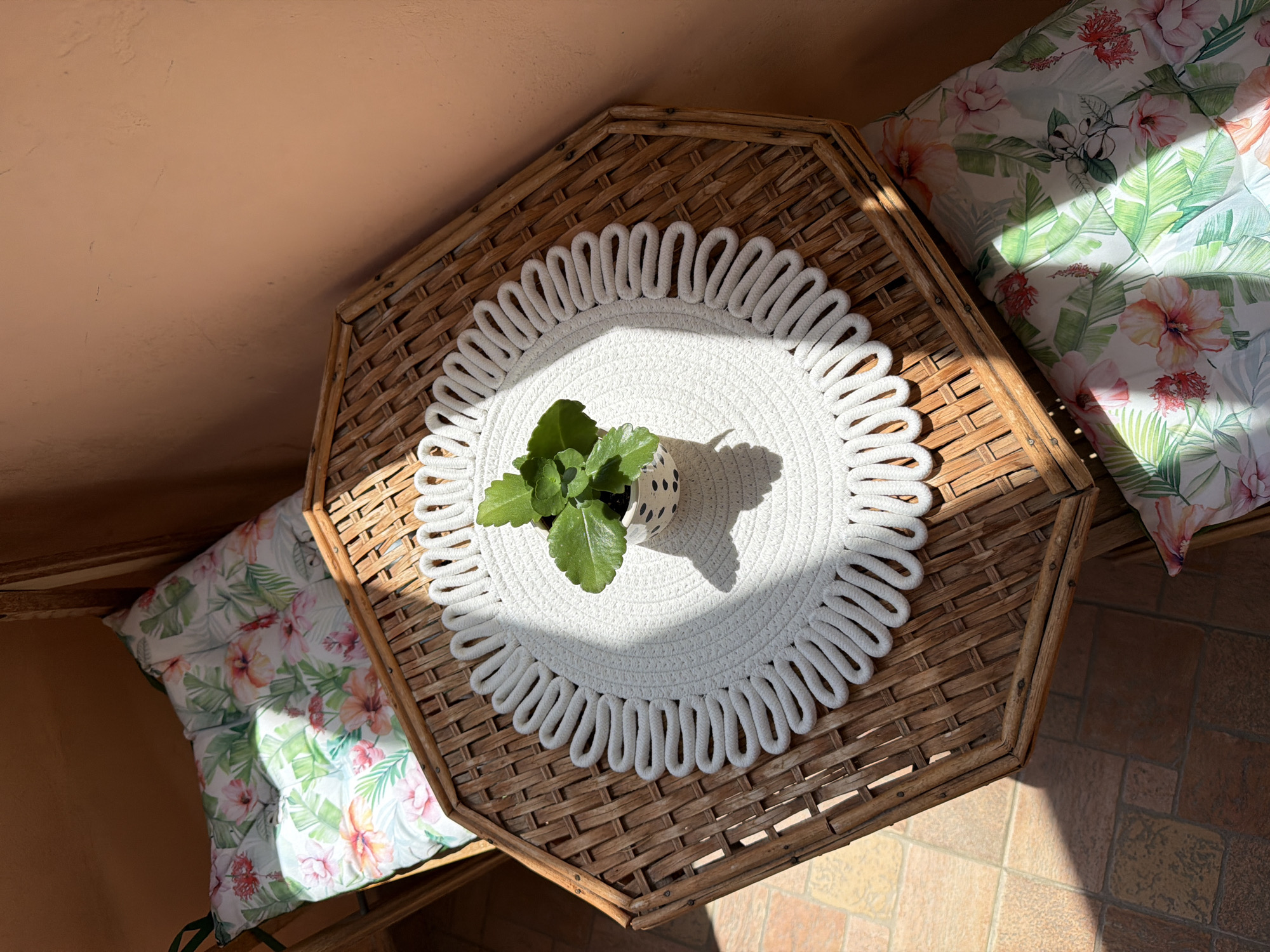This overhead shot captures a tranquil corner, likely a sunroom or covered patio in Lovech, Bulgaria, illuminated by direct sunlight. At the center of the frame is an octagonal wicker table with a tightly woven texture. A round, white placemat, resembling macrame with a distinctive looped border, is centrally placed on the table. Resting on the placemat is a small, vibrant green plant, possibly a succulent, potted in a white ceramic container adorned with black polka dots.
Strong, angular shadows cast by the plant, pot, and table indicate bright sunlight, suggesting either morning or late afternoon. The light creates a dynamic contrast between illuminated surfaces and shadowed areas across the scene.
To the upper right and lower left, portions of two plush cushions are visible, featuring a lively tropical floral pattern with large pink and orange hibiscus flowers and lush green leaves. These cushions suggest a comfortable seating arrangement next to the table. The upper left corner reveals a plain, textured wall or ceiling in a warm, light terracotta or peach color. The floor in the lower right consists of irregularly shaped, earthy-toned tiles in shades of brown and beige. No individuals are present, and no text is visible in the image, contributing to a serene and inviting ambiance.