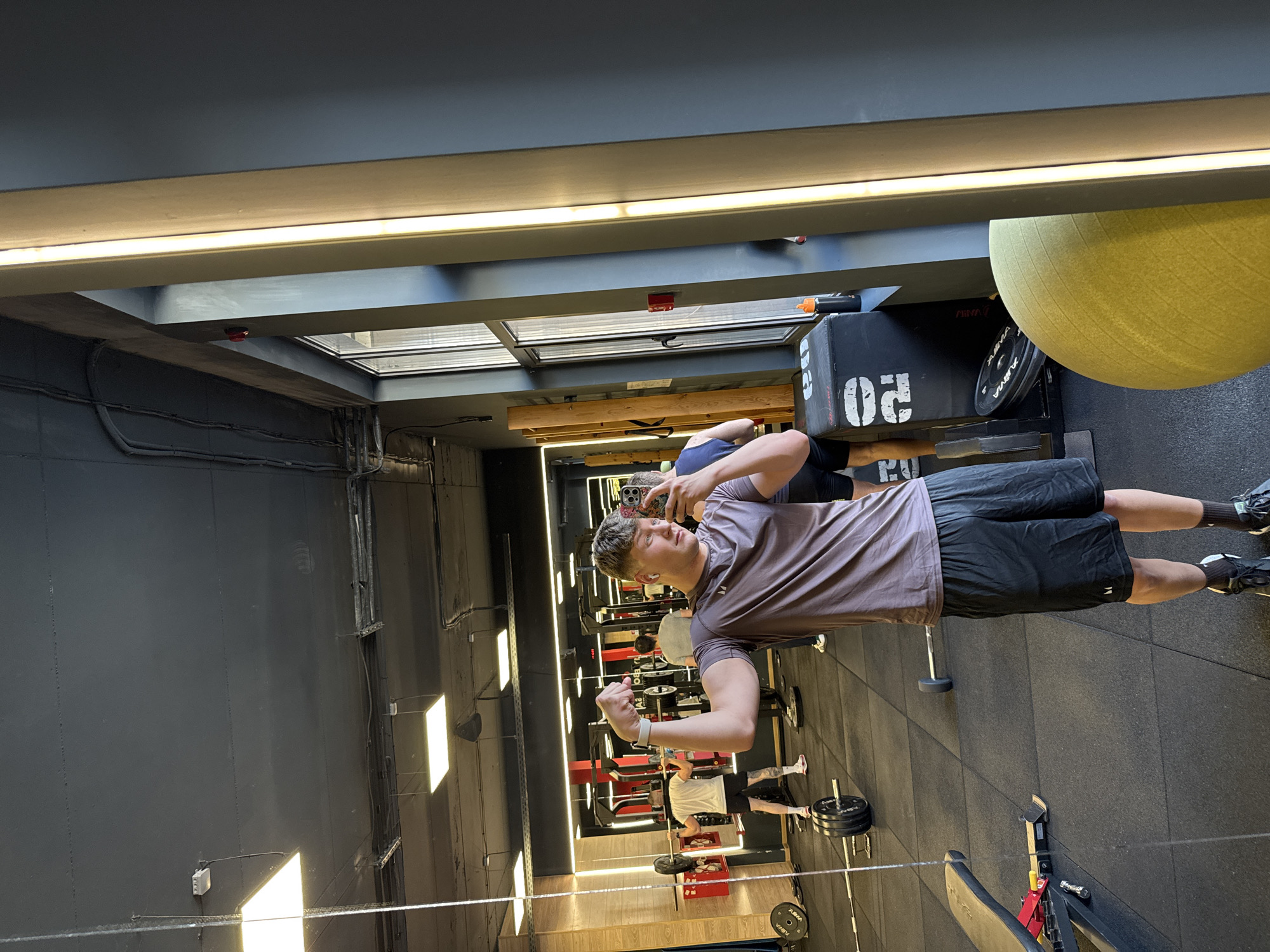 This image depicts an indoor gym located in Plovdiv, Bulgaria, during what appears to be daytime, given the natural light entering through overhead skylights or large windows. The gym features dark gray walls and a ceiling structure with exposed beams and linear LED lighting, creating a modern and functional atmosphere.
The primary subject is a young man, likely in his late teens or early twenties, who is capturing a mirror selfie. He has light brown hair and is dressed in a short-sleeved, collared gray/brown t-shirt and black shorts. He flexes his left bicep, proudly displaying a white wristband on his left wrist, while holding a smartphone with a colorful, patterned case in his right hand to take the picture. His expression suggests confidence and satisfaction, possibly reflecting a successful workout. Earbuds are visible in his ears.
In the background reflections, other gym patrons are partially visible, including one person in a white t-shirt and black shorts, engaged with equipment. The gym is well-equipped with various apparatuses: black plyometric boxes, one marked with "02," weight plates, some branded "ALLMA" and one with "20," barbells, and a yellow-green exercise ball. A red and black weight bench is also seen in the foreground. Overhead, a red fire alarm device is mounted on a beam, and electrical conduits run along the walls, alongside a white electrical box. The floor is covered in dark rubber gym matting.