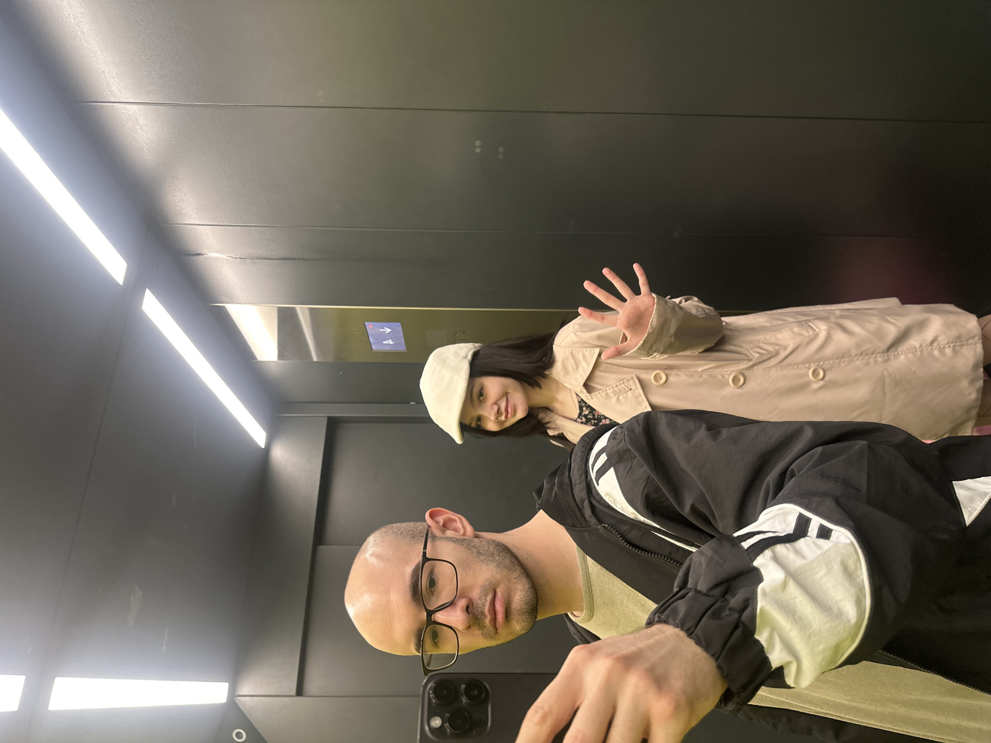 A selfie of two people inside an elevator. The man taking the selfie is bald, wearing glasses, and a black and white jacket. He is holding a black smartphone with a multi-lens camera. Behind him, a woman with dark hair is wearing a white cap and a beige trench coat. She is waving her hand. The elevator has fluorescent lighting and dark gray paneled walls. A panel with elevator direction arrows is visible. The location of the photograph is Lyubertsy, Russia.