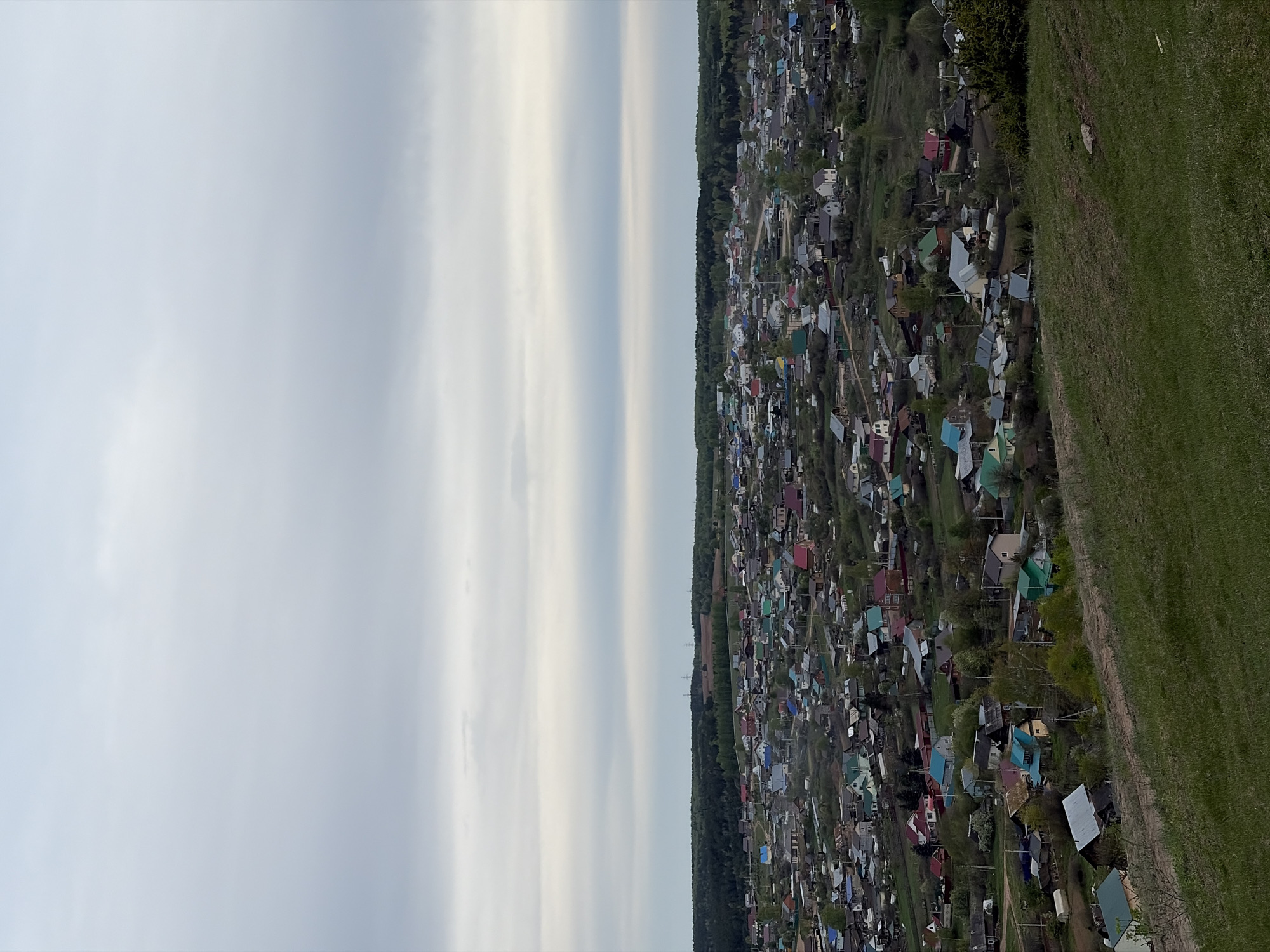 This aerial shot shows a rural part of Ольховка, Russia. The left portion of the image is dominated by a gray sky with some cloud formations. The right side of the image shows a dense cluster of residential buildings with colorful roofs surrounded by greenery. A grassy area extends from the edge of the built-up area.