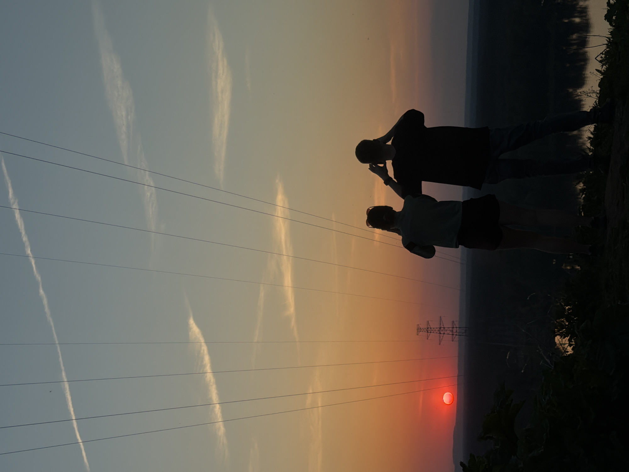 At sunset in Noviy, Russia, two people stand silhouetted against the orange-red sky, near an electric transmission tower. Power lines stretch across the sky. One person is holding a mobile phone up to their face.