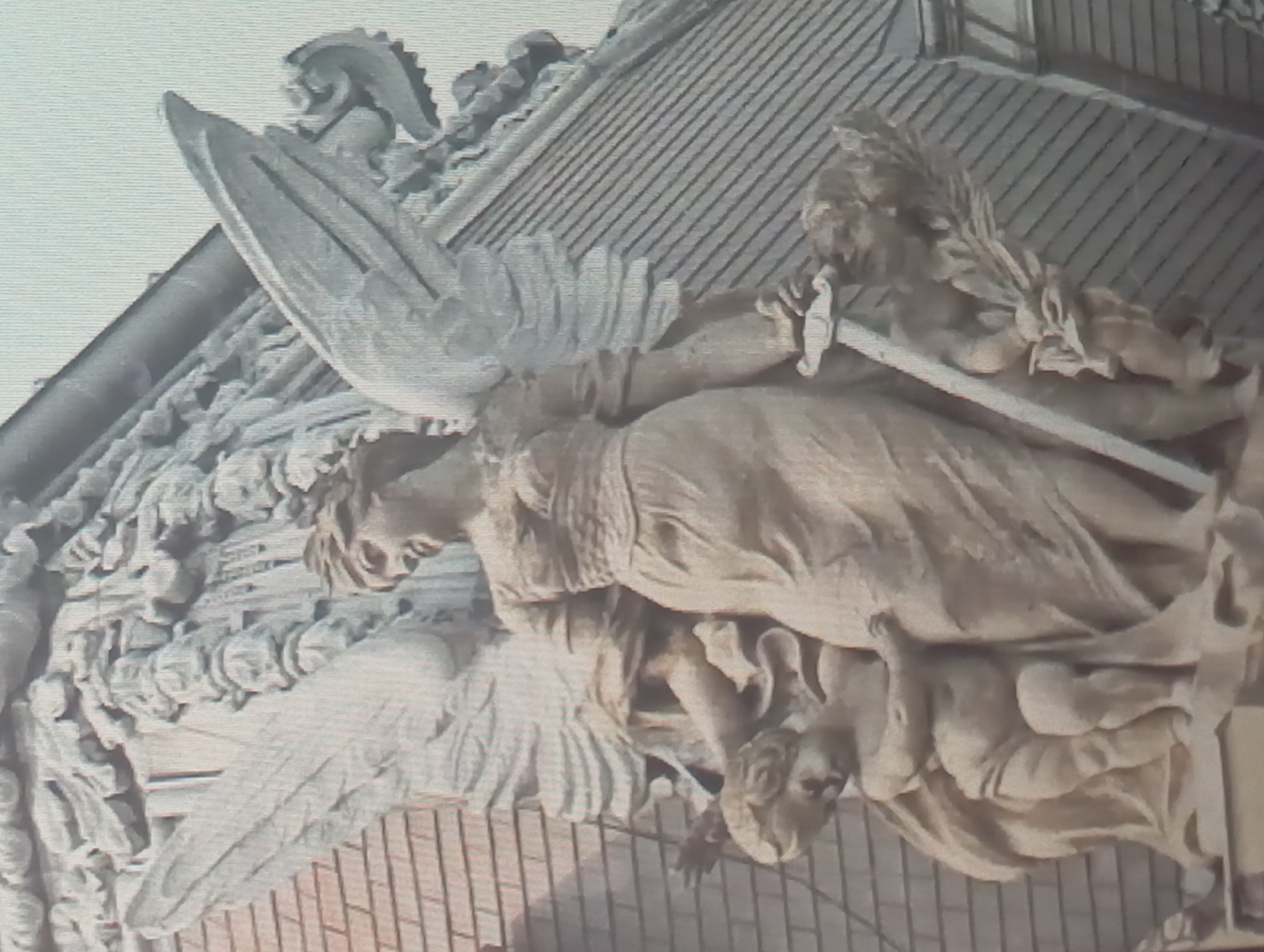 The image depicts a highly detailed stone sculpture of an angel figure, possibly a statue or architectural embellishment. The angel possesses large wings, draped clothing, and appears to be holding a long, straight object, which might be a sword or staff. Behind the angel figure, there is another sculpted figure or part of a building with decorative elements. The background shows what appears to be a building roof or a wall with a repetitive pattern.
Given the request context, this sculpture might be part of a building in Karaganda, Kazakhstan, but without further visual cues or information, it is impossible to pinpoint its exact location.