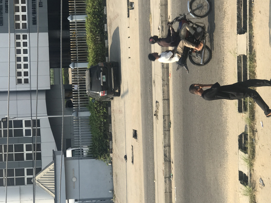 This image depicts an urban street scene in front of the Christopher Ola High/Magistrate Court, Eti-Osa Local Government Area, Lagos State, Nigeria. The background features a modern, multi-story building with a grey facade and numerous windows, identified by visible text reading "CHRISTOPHER OLA COURT HOUSE HIGH/MAGISTRATE COURT ETI-". The court building is secured by a grey metal fence, behind which a dark-colored SUV is parked on a paved lot. A smaller grey utility building stands near the fence line on the left. In the middle ground, a multi-lane paved road with a concrete curb separates the courthouse property from a dirt and sparsely vegetated shoulder. Two males are observed riding a red and silver motorcycle from right to left on the road; the driver wears a dark cap and shirt, while the passenger wears a white t-shirt and appears barefoot. In the immediate foreground, a male pedestrian dressed in a dark tunic and pants walks leftward on the dirt shoulder, his right hand raised to his ear. The scene is illuminated by direct daylight, casting clear shadows. The road and shoulders exhibit minor debris and surface imperfections, including open depressions in the paved pedestrian areas.