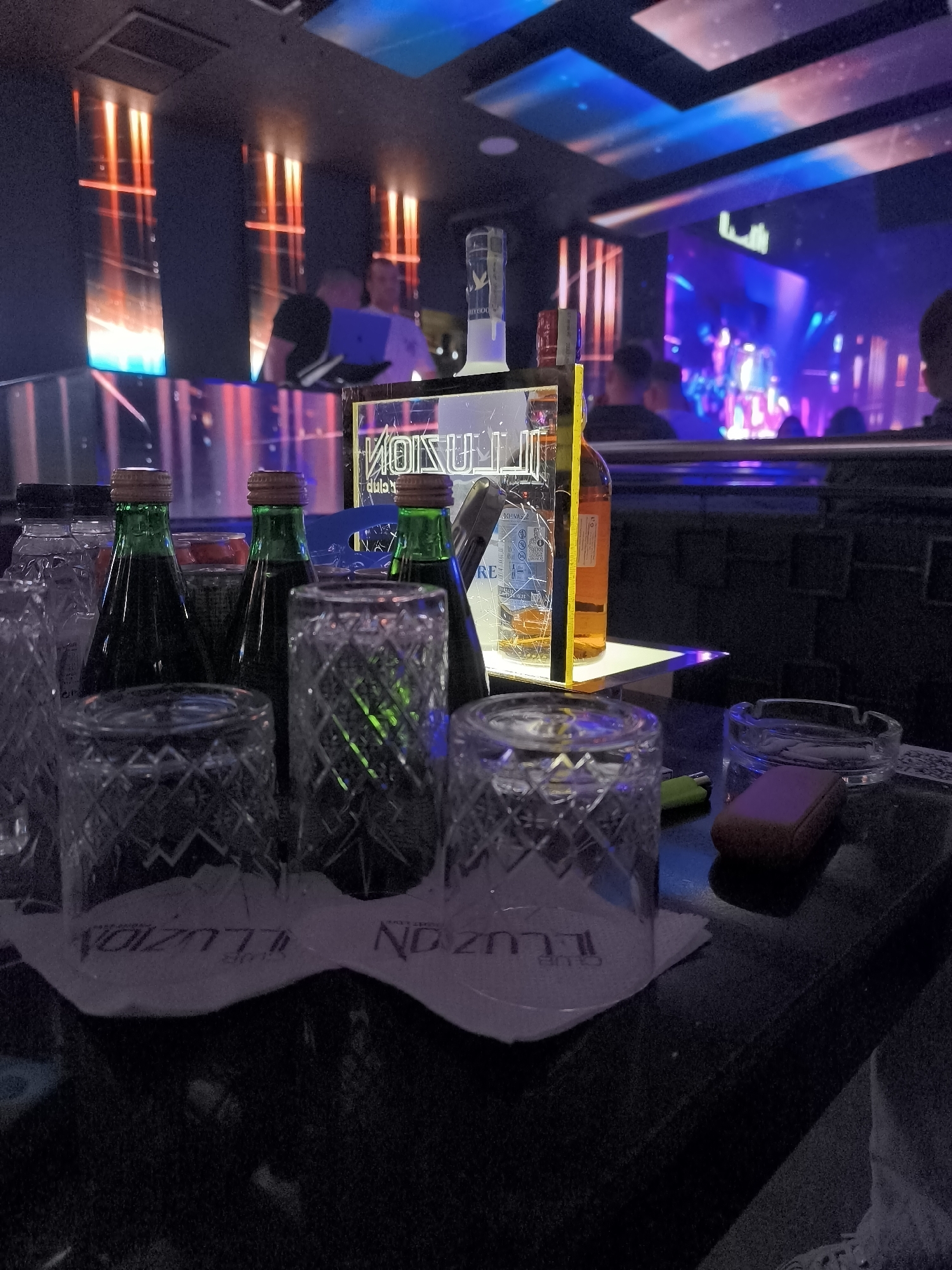 The photograph captures a scene inside a nightclub, likely located in Sofia, Bulgaria, given the presence of branded items with the name "CLUB ILUZION." The immediate foreground features a table setting with ornate glass tumblers placed on top of napkins bearing the club's name. Also visible are bottles of liquor, and an ashtray. Patrons are visible in the background. The overall ambiance suggests a dimly lit, upscale nightlife setting with colorful lighting.