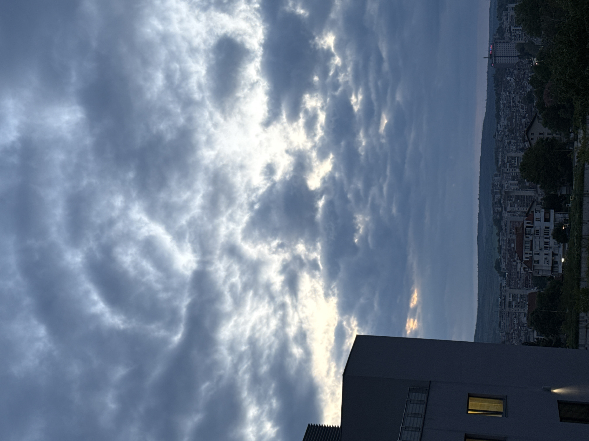 The image shows a cloudy sky over Pleven, Bulgaria. The low angle suggests the picture was taken from a lower vantage point, possibly a building. The city of Pleven is visible in the background, with multiple buildings and a hilly landscape. The sky is dominated by gray clouds, with some patches of lighter areas indicating the sun's position. A corner of a building is in the foreground. The buildings' lights are on.