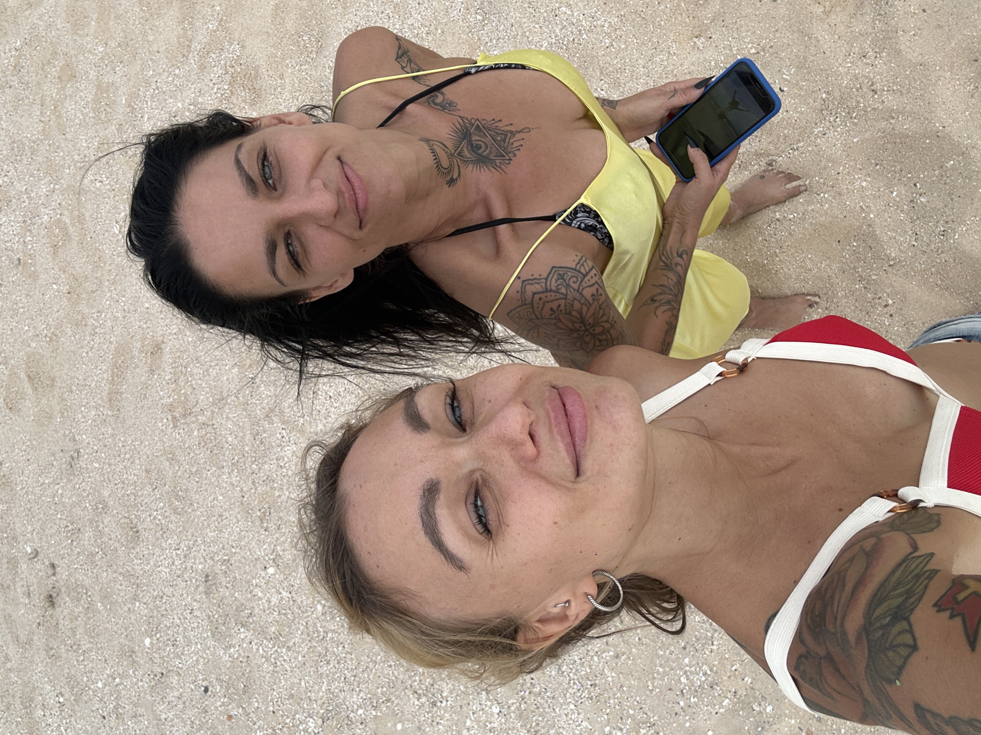 Two women are lying on a sandy beach. The woman at the top of the image has dark hair and is wearing a yellow dress with black straps. She is holding a phone with a blue case. The woman at the bottom of the image has blonde hair and is wearing a red and white bathing suit. Both women have tattoos. The location is Phu Thuy Ward, Vietnam.