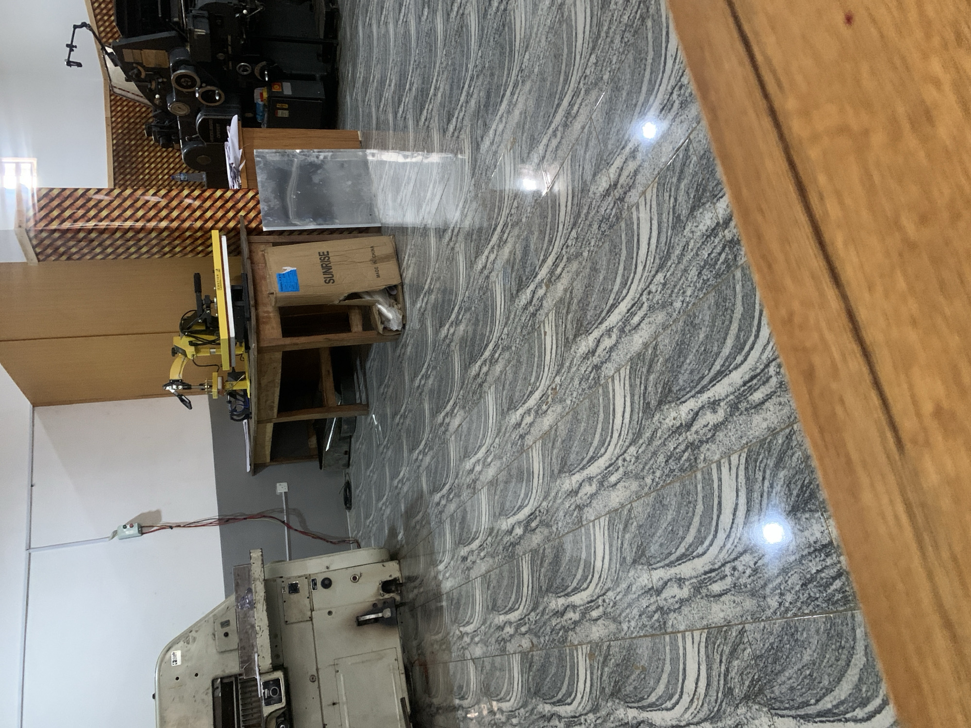 The image depicts an indoor scene, possibly a workshop or printing facility. In the foreground, a section of a wooden table or counter is visible. The floor is tiled with a marbled pattern in shades of grey and white, exhibiting a high-gloss finish that reflects light.
Dominating the left side of the image is a large, beige industrial machine, likely a paper cutter or printing press, with visible mechanical components and control panels. To the right of this machine, a wooden table supports a yellow heat press machine with a black arm. Next to the table is a cardboard box labeled "SUNRISE" with "MADE IN CHINA" printed below.
Further to the right, a large, black industrial printing press is partially visible, characterized by its complex machinery and numerous drums. A clear acrylic or glass sheet stands upright near the printing press. The walls are painted in a neutral tone, with a section of orange and yellow striped wallpaper visible on a pillar. Electrical conduits and a junction box are mounted on the wall. The overall impression is one of a functional industrial space.