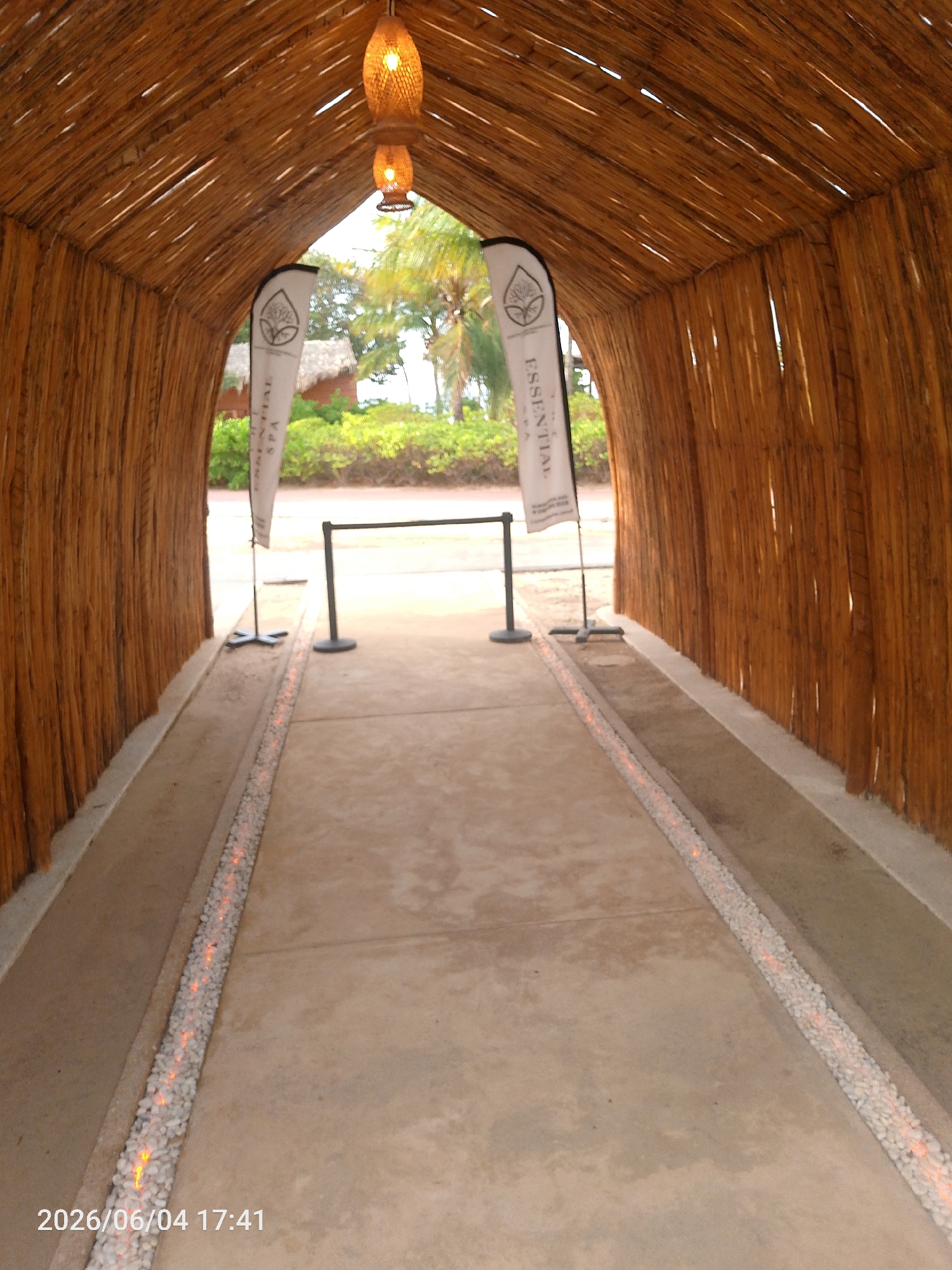 The image captures a unique, tunnel-like pathway constructed from numerous light-brown wooden poles or bamboo, forming an arched ceiling and vertical walls. Inside this structure, two woven, basket-like pendant lamps hang, emitting a warm, orange light.
A light-colored concrete path extends through the tunnel, bordered on both sides by narrow channels filled with small white pebbles. These pebble channels feature embedded orange lights, creating an illuminated track along the walkway.
Towards the exit of the tunnel, two white banner flags are prominently displayed. Both flags feature a logo of a stylized tree or plant within a drop shape, and the vertical text "ESSENTIAL SPA." Two black stanchions with round bases are placed across the path, suggesting a controlled entry point.
Beyond the tunnel, a bright outdoor scene is visible, featuring lush green bushes, tall palm trees, and a building with a thatched roof, indicating a tropical or resort-like environment. No individuals are present in the image. The warm interior lighting and the natural materials evoke a serene and inviting atmosphere, likely for a spa or wellness center. The timestamp "2026/06/04 17:41" is visible in the bottom left corner.