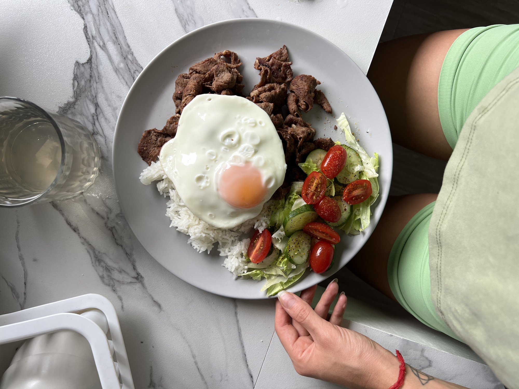 A high-angle shot depicts a plate of food on a marble surface, with a partial view of a person in the scene.
The plate is a light gray color and contains a portion of white rice topped with a fried egg, pieces of cooked beef, and a small salad consisting of lettuce, halved cherry tomatoes, and cucumber slices. A hand is visible holding the edge of the plate.
To the left of the plate, there is a glass filled with liquid. A white container is also partially visible in the lower left corner of the image.
The person is wearing light green shorts and a light green t-shirt. A red bracelet is on the person's left wrist.
The scene appears to be indoors, on a marble countertop, and the location is Unknown, Unknown.