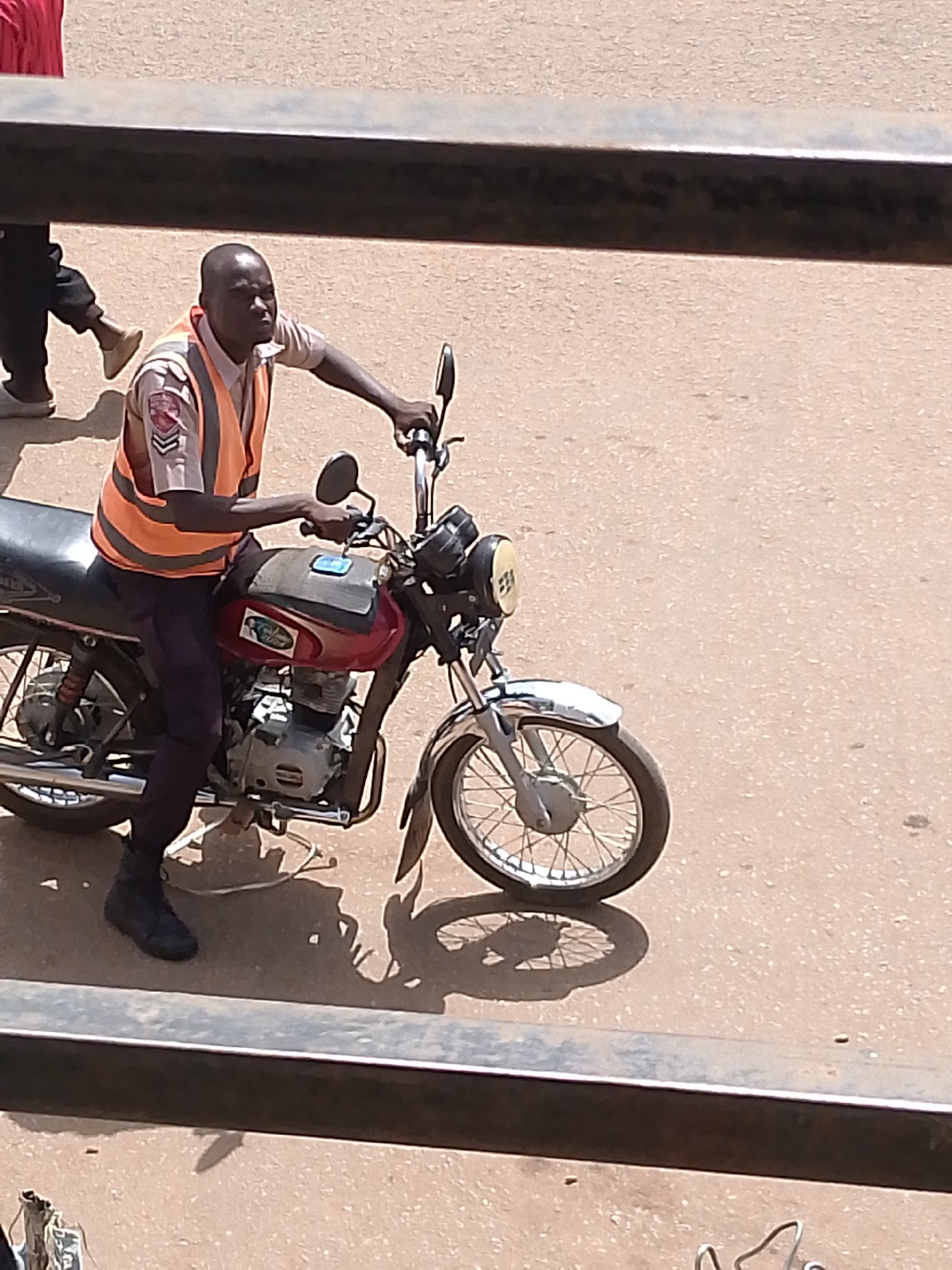 In Bukuru, Nigeria, a man wearing an orange safety vest over a light-colored shirt and dark pants sits astride a red motorcycle. The motorcycle has a chrome front fender and spoked wheels. The man's left hand is on the handlebar. The scene is viewed from an elevated position, with a dark metal bar in the foreground obscuring the bottom portion of the image. The ground appears to be a dusty road.