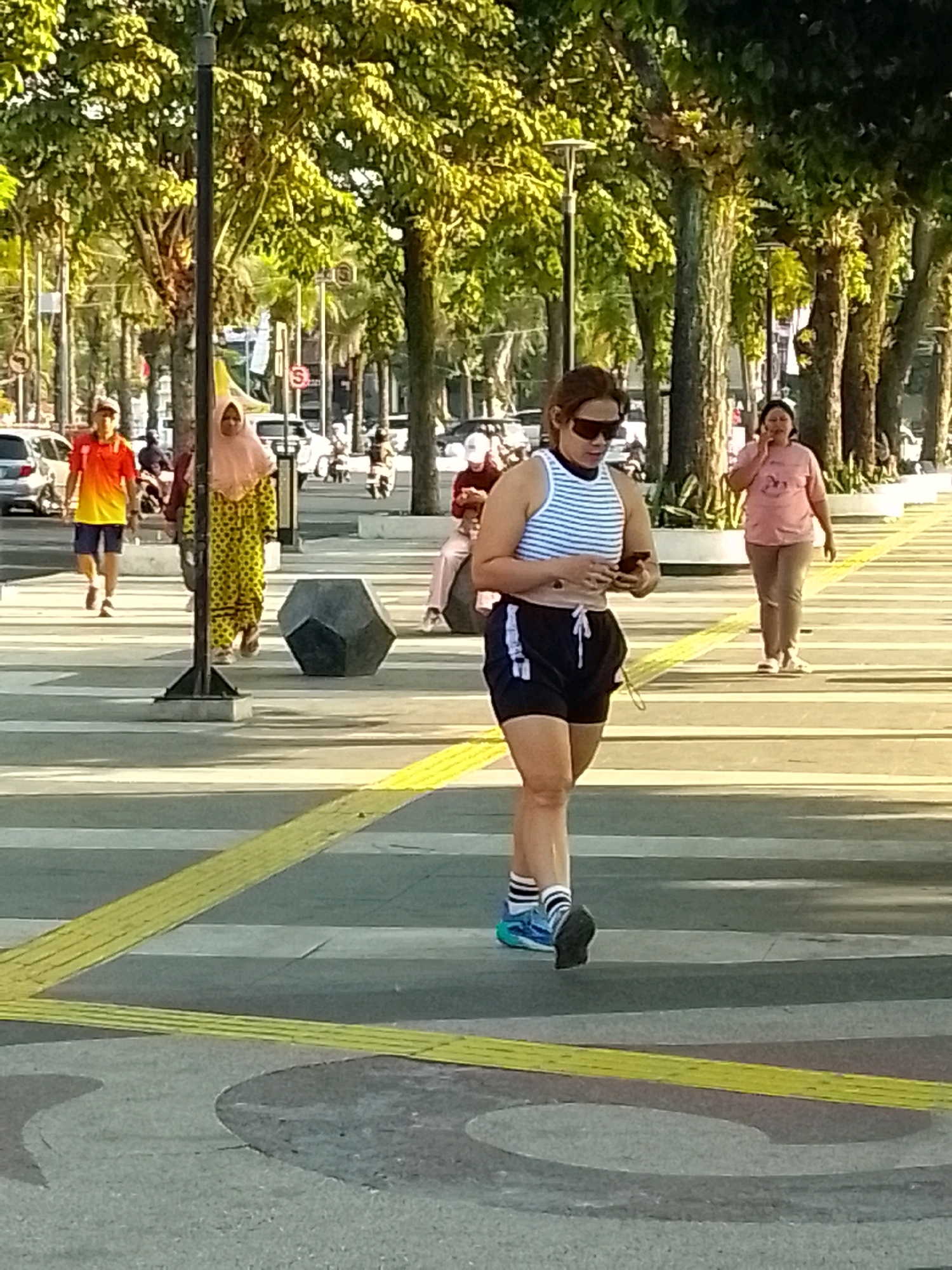 This is a ground-level, eye-level, medium shot of a pedestrian walkway in Jember, Indonesia, possibly along Jalan Gajah Mada. The sun is casting long shadows from the trees lining the walkway. In the foreground, a woman with dark hair, wearing sunglasses, a white and blue striped tank top, and black athletic shorts is walking forward and looking down at her phone. She has on blue and white athletic shoes and striped socks. Behind her and slightly to her left, a person wearing a bright yellow and orange gradient t-shirt and dark shorts is walking away from the camera. To the left of that person, a woman in a yellow and black polka-dot dress and a light pink hijab is walking towards the camera. Further back and to the right, a woman wearing a pink t-shirt and beige pants is talking on her phone. The walkway is paved with light-colored concrete with yellow tactile paving strips. In the background, cars are visible on a street, and more trees and foliage line the walkway. A large, dark, polygonal concrete sculpture is positioned on the walkway between the main subjects. The overall impression is of a bright, sunny day in a public urban space.
