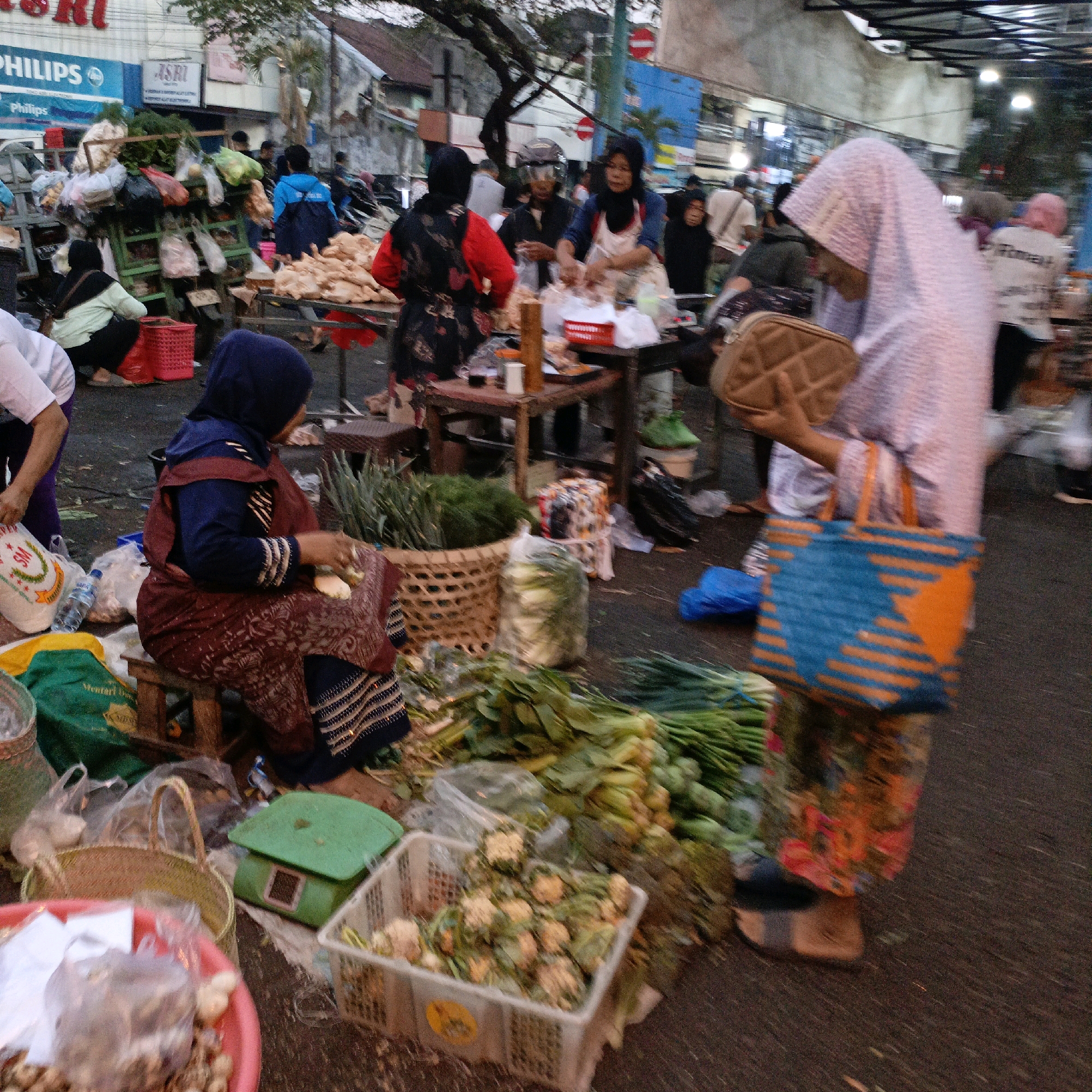 The image depicts an outdoor marketplace in Salatiga, Indonesia. In the foreground, a woman in a dark blue hijab and maroon and black patterned clothing sits on a small wooden stool. She is holding a piece of white food in her right hand and appears to be preparing it, with her left hand resting on a basket. To her left, a large woven basket sits on the ground. In front of her, there is a pink plastic basin containing more food items, some of which are in plastic bags. A green scale is also visible to the left of the basket.
Further into the background, a variety of produce, including leafy green vegetables and what appears to be cauliflower, is displayed. People are engaged in various activities, such as tending to stalls, shopping, and conversing. Some individuals are wearing hijabs, consistent with the local culture. The environment suggests a busy market setting with stalls and goods laid out on the ground and tables. The lighting indicates it is either early morning or late afternoon, with a somewhat dim and overcast atmosphere. The overall scene captures the essence of a local Indonesian market.