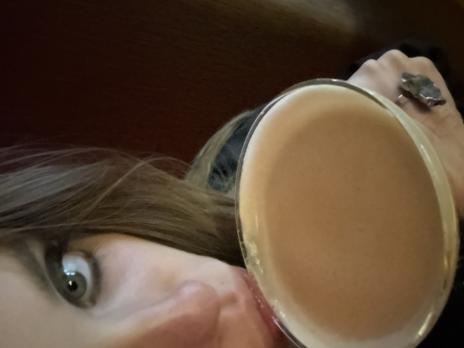 A person's face is tilted to the left, with a single, wide eye visible. Brown hair falls across the forehead. A cocktail glass dominates the foreground, filled with a creamy, light brown drink. A hand holding the glass is visible, adorned with a silver ring featuring a large, irregularly shaped stone. The background is out of focus, with a dark red surface and dark clothing.