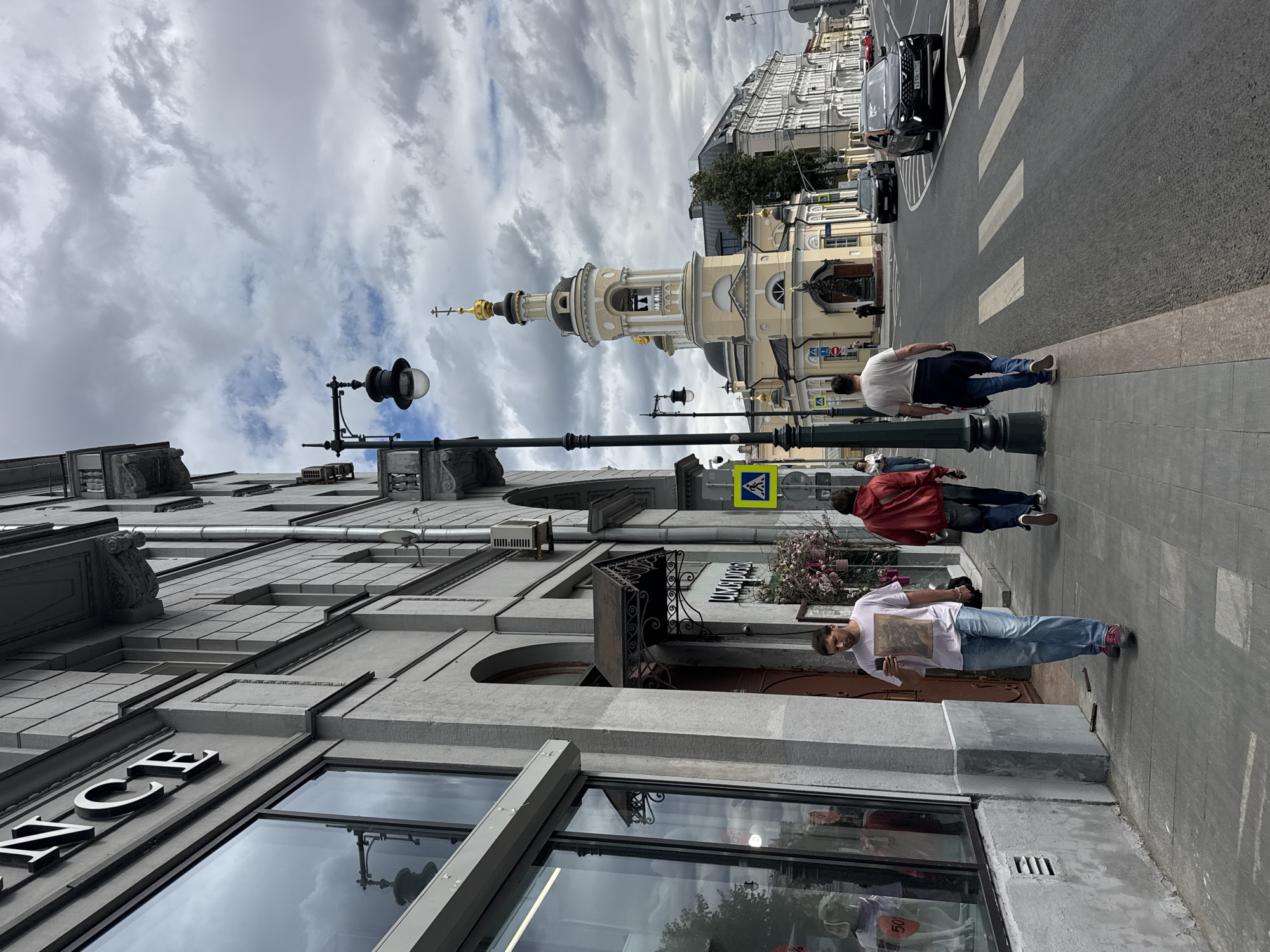 This is a street scene in Moscow, Russia, on a cloudy day. The perspective is from a sidewalk looking down a street. On the left, a grey building with large windows is visible, with the partial word "NCE" in large, raised letters on its facade. The windows reflect the cloudy sky and some street lamps. A few people are walking on the sidewalk. In the foreground, a man wearing a light-colored t-shirt and blue jeans is walking while looking at his phone. Further down the sidewalk, another man in a red jacket and blue jeans walks away from the camera. Across the street, a distinctive yellow church with a golden dome and bell tower stands prominently. A statue is located in front of the church. Cars are visible on the road, including a black SUV and a dark sedan. Pedestrian crossings are marked on the asphalt. The overall environment appears to be a mix of commercial and religious architecture.
