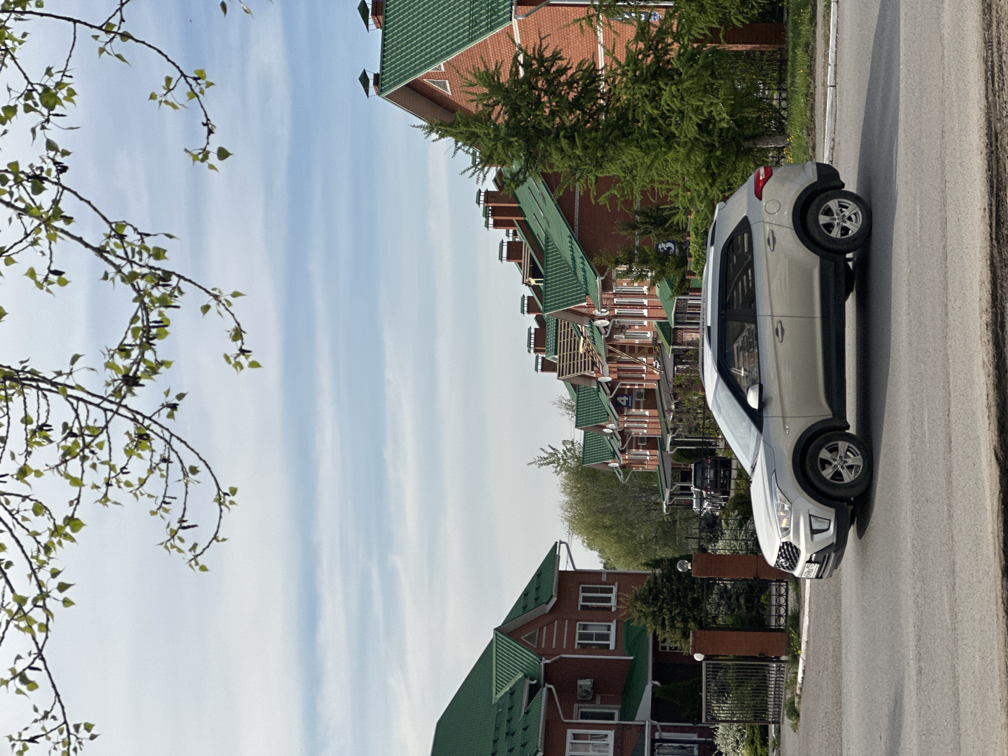 The image depicts a street scene in the city of Chaikovsky, Russia. A silver Hyundai Creta is parked on the right side of a paved road. A series of multi-story residential buildings are visible on the left side of the road. The buildings are characterized by red brick exteriors and distinctive green, tiled roofs. A tree branch with budding leaves occupies the left foreground of the image. The sky is partly cloudy.
