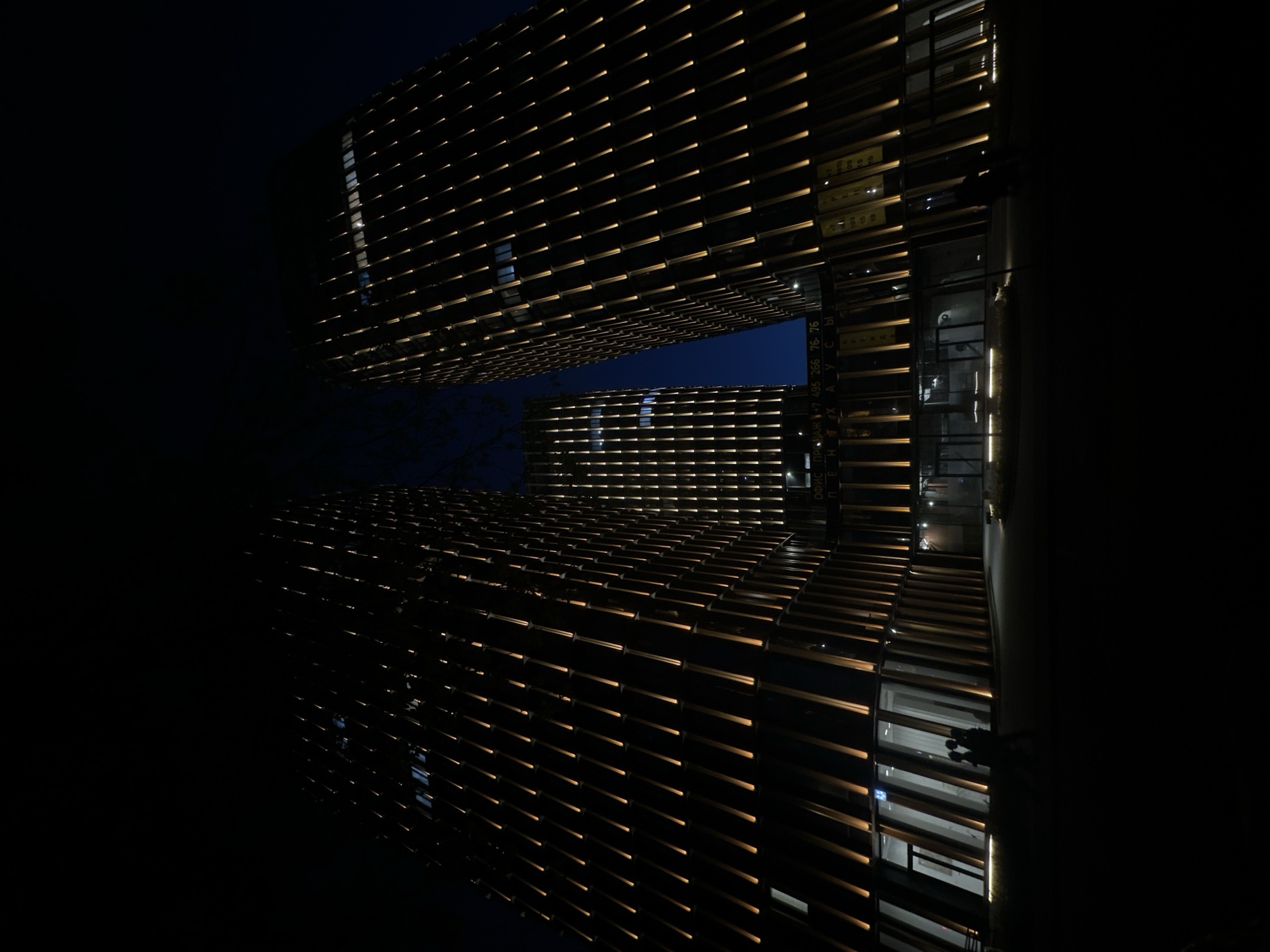 The image shows the exterior of a modern building in Moscow, Russia, at night. The building features a distinctive facade with horizontal illuminated slats. The overall lighting is low, emphasizing the architectural details and creating a contrast with the dark sky. A second building is partially visible in the background. The image is taken from a low angle, looking up at the buildings.