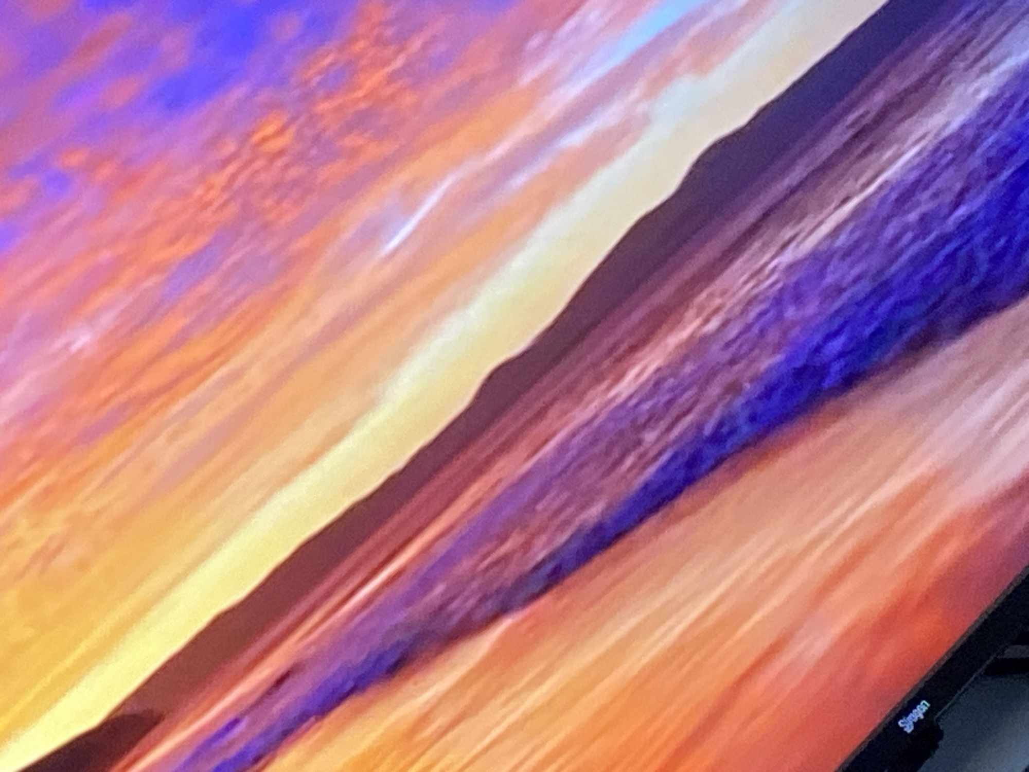 The image is a close-up shot of a computer monitor displaying a colorful abstract image. The colors present include orange, yellow, white, blue, and purple, seemingly blended together in a painterly fashion. In the bottom right corner, a portion of the monitor's frame and a "Siragon" brand label are visible, indicating the manufacturer. The abstract image is likely a background or screensaver on the monitor. No specific location in Cotoperíz, Venezuela can be determined as the image is of the monitor display. No people, subjects, actions, or interactions can be described.