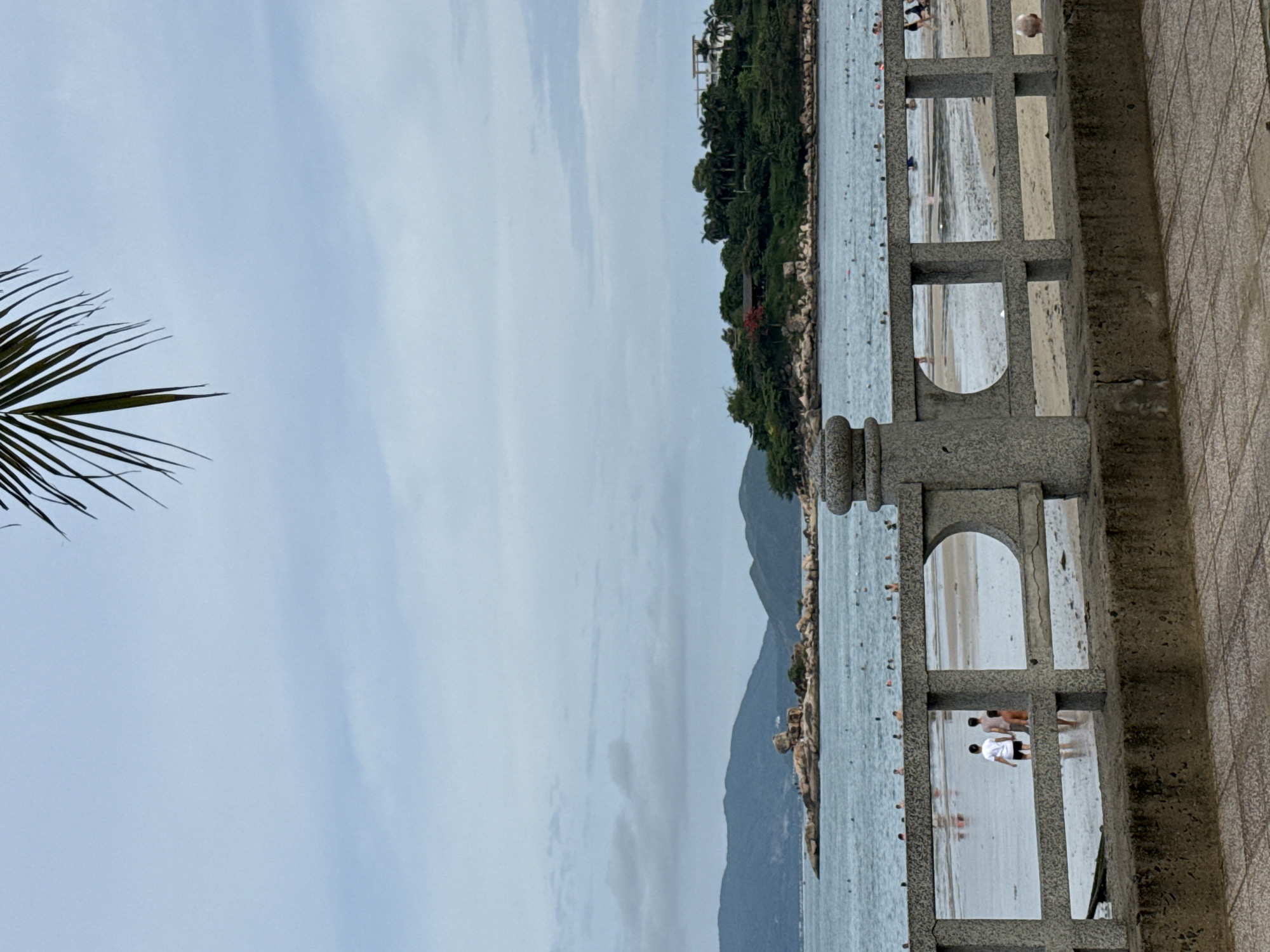 The image displays a tranquil coastal scene, likely a public beach or promenade in Bac Nha Trang Ward, Vietnam, under an overcast or partly cloudy sky. In the foreground, a sturdy grey stone railing, featuring both square and arched openings, runs diagonally across the lower right portion of the frame. To the right of the railing, a textured grey pavement or walkway is visible.
Beyond the railing, a calm, light blue sea stretches towards a verdant, forested landmass in the background. This distant coastline is characterized by dense tree cover, hills, and mountains, with a few buildings, possibly resort structures, visible on the higher slopes. Rocky outcrops punctuate the shoreline.
Numerous people are enjoying the water and the sandy areas. Through one of the railing's arched openings, two individuals are clearly visible standing on a light-colored sandy stretch or in very shallow water: one in a white t-shirt and dark shorts, and another in a light-colored swimsuit. Many other figures appear as small, blurred forms scattered across the sea, suggesting swimming or wading. A dark green palm frond partially frames the pale blue-grey sky in the upper-left corner, adding a tropical touch. The diffused daylight indicates it is daytime, likely late morning or afternoon. There is no visible text in the image.