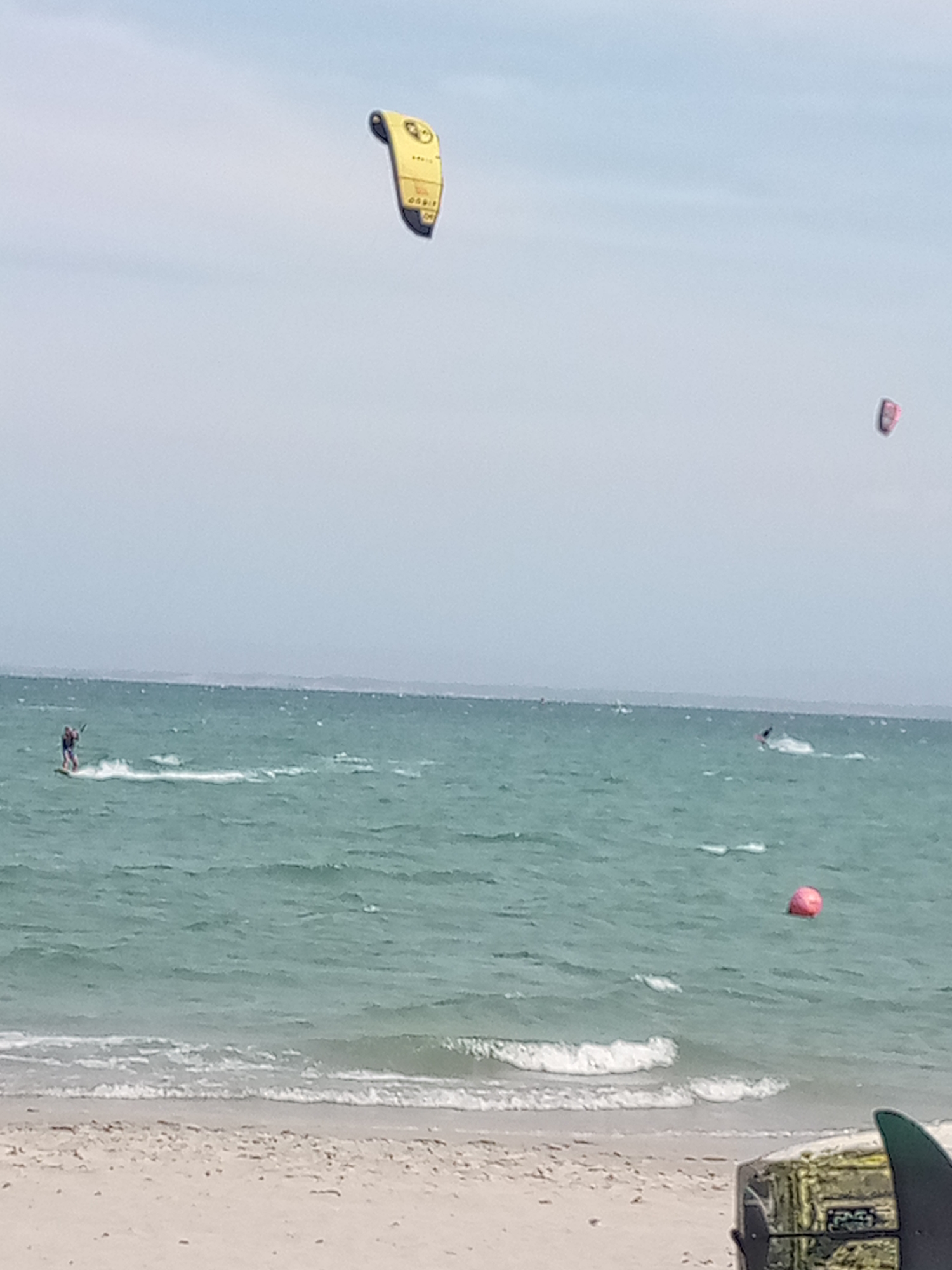 The image depicts a coastal scene in El Yaque, Venezuela, featuring kitesurfing activity on the water. Two kites are visible in the sky: one yellow and one pink. There are two kitesurfers and one buoy visible in the water. The beach is sandy, with gentle waves approaching the shore. In the bottom right corner of the frame, a partially obscured box-like object and a fin are visible. The sky is overcast.