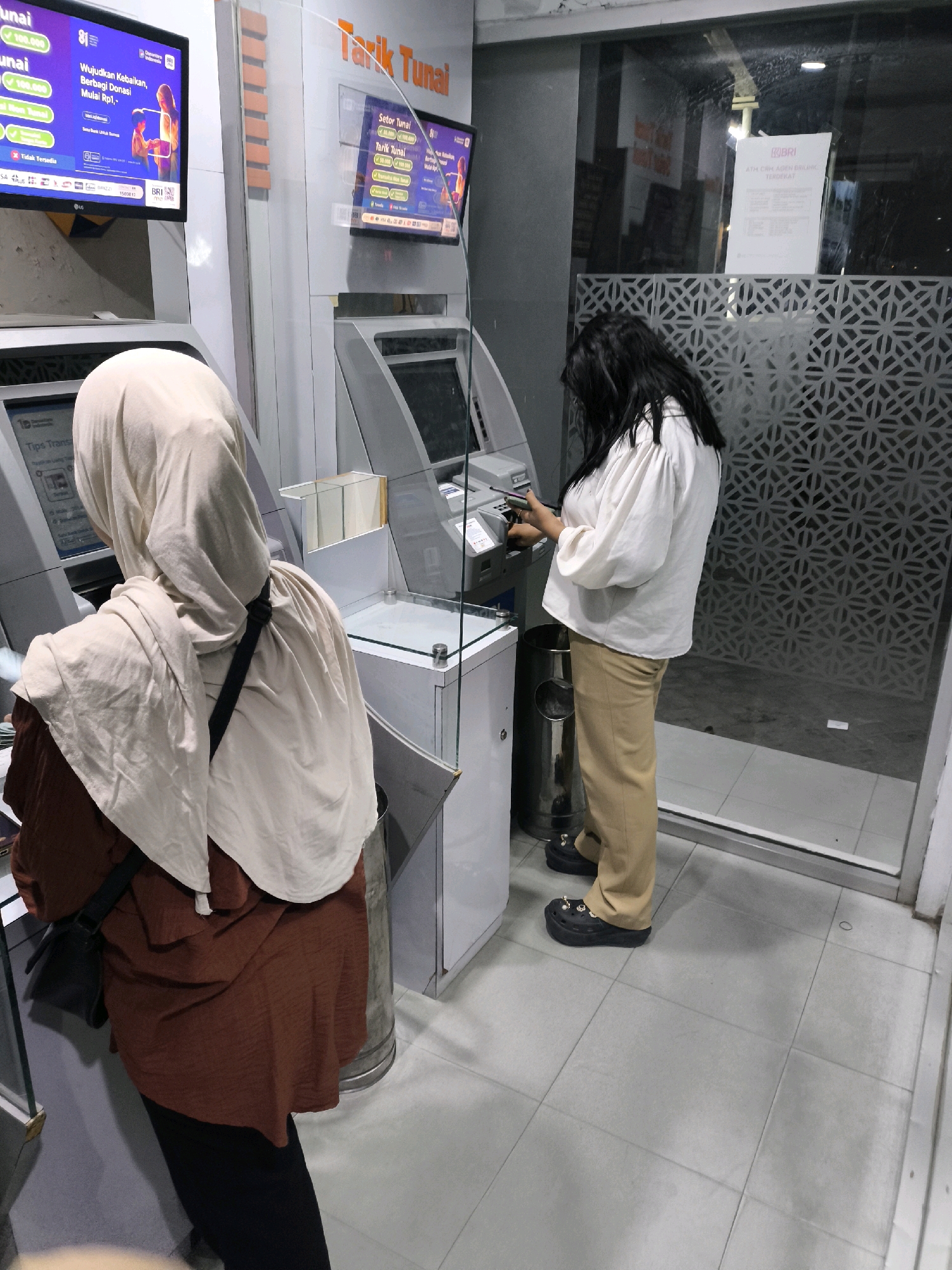 In Limboto, Indonesia, two individuals are captured in a scene at an ATM. The first individual, wearing a brown tunic and a beige hijab, is positioned in front of an ATM screen, their back to the camera. They are holding what appears to be a card or phone. Beside them, a metallic trash receptacle is visible.
The second individual, standing to the right, is facing an ATM machine, also with their back partially to the viewer. This person is wearing a white blouse with puffed sleeves and tan pants. They are actively engaged with the ATM, holding a smartphone in their hands. A large metallic trash receptacle is also present next to them.
The environment suggests an indoor setting, possibly a bank or a commercial establishment, with tiled flooring and glass partitions. The ATM screens display various transaction options and promotional content. The overall lighting is artificial, indicating it is likely evening or a dimly lit interior space. The glass partition features a patterned design. A sign is affixed to the glass with text that includes "ATM AGEN BRI..." suggesting a banking service point.