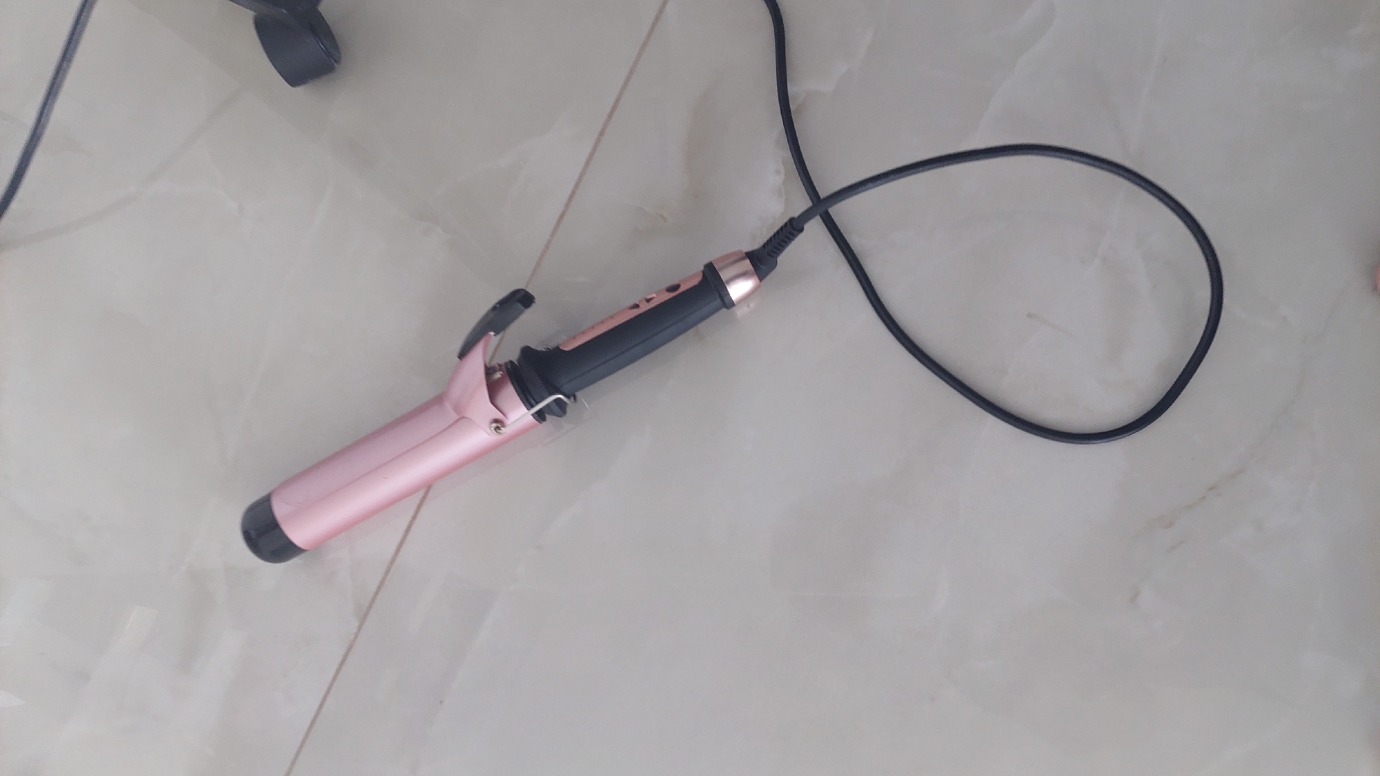A pink and black curling iron with a black power cord is lying on a light-colored tile floor. The environment appears to be indoors.