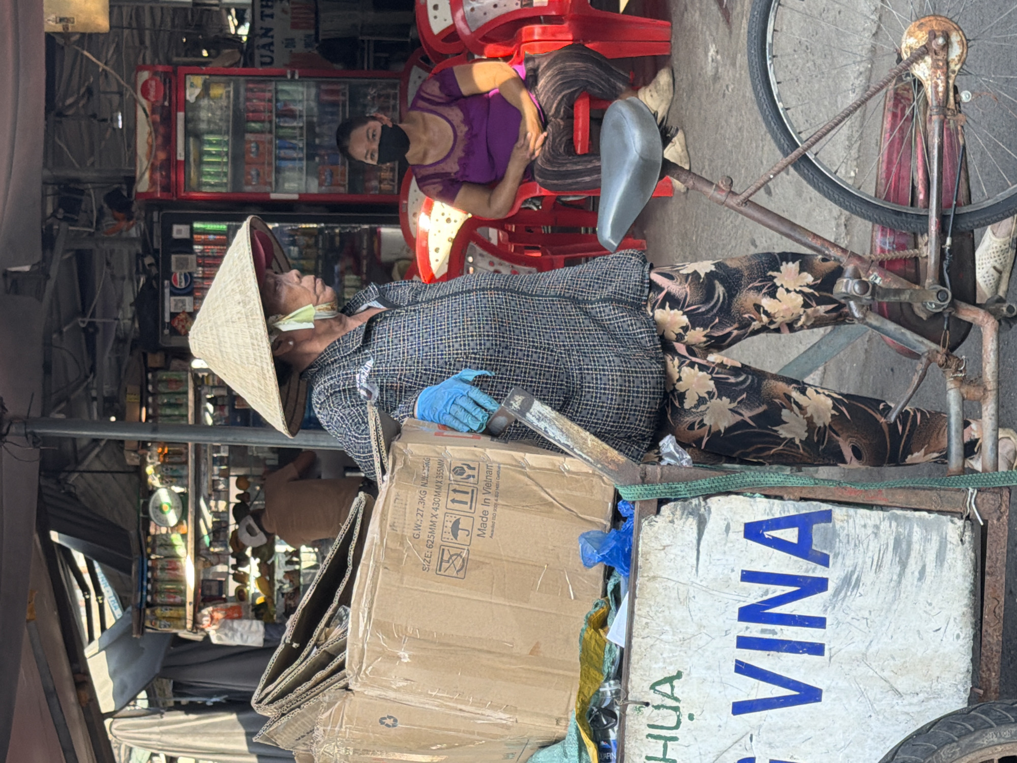 In Nha Trang, Vietnam, an elderly woman wearing a conical hat and a checkered shirt stands next to a cart piled with cardboard boxes. She has a light green cloth tied around her face, and her hands are covered with blue gloves. The cart is branded with the words "G VINA" in large blue letters. A woman in a purple shirt and face mask sits on a red plastic chair in the background. Refrigerated drink coolers and shelves stocked with goods are visible behind the people. The scene appears to be an outdoor market or roadside stall.