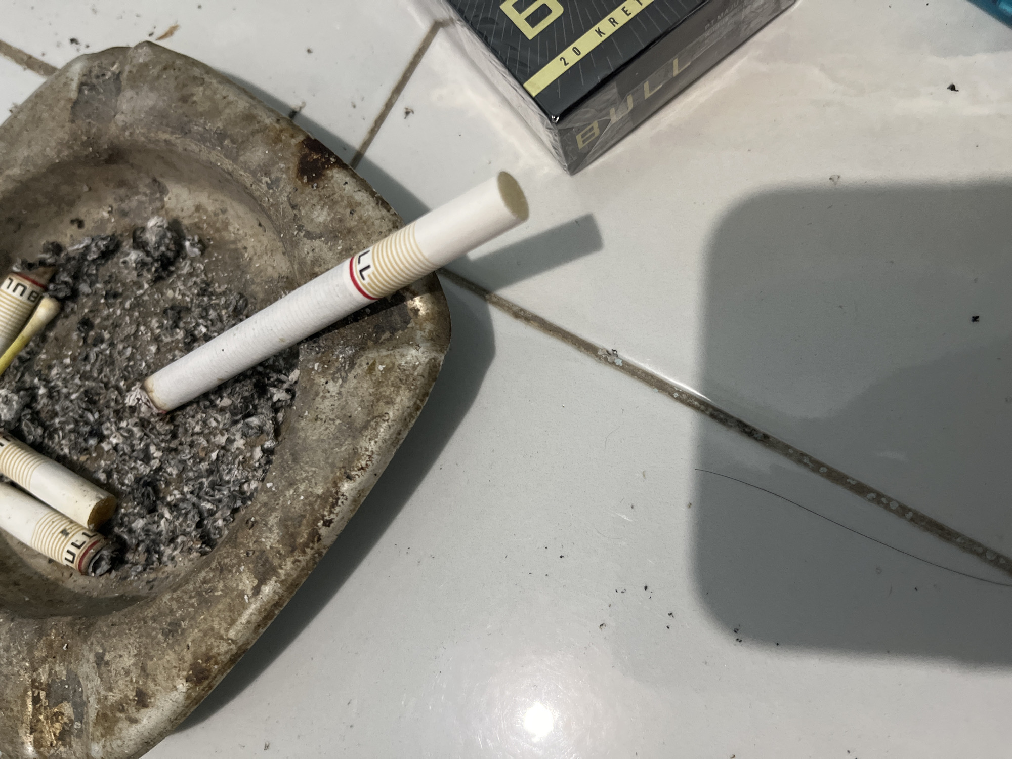 In an indoor setting, likely a tiled floor, a scene involving cigarettes is depicted. An ashtray, appearing weathered and metallic, is filled with cigarette ash and several cigarette butts. One cigarette lies partially inside the ashtray, while another is positioned outside, resting on the floor. To the right of the ashtray, a partially visible pack of cigarettes is present. The overall composition suggests a moment of rest or a break, with the presence of the cigarettes and ash indicating recent use. The location is identified as Sumbernadi, Indonesia.