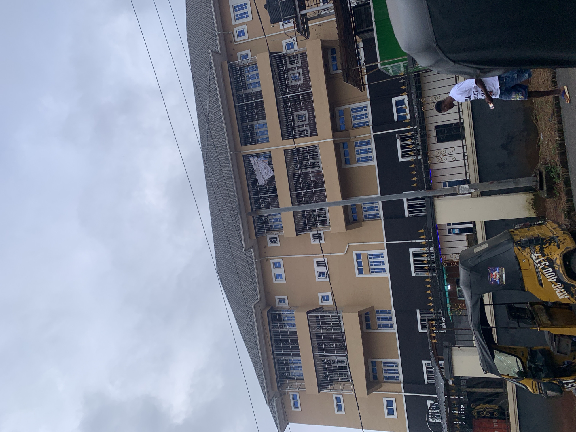 In the city of Nkpor, Nigeria, an overcast sky looms above a multi-story building. The building is painted in two tones: a light beige on the upper floors and a dark charcoal gray on the lower sections. Each floor features numerous windows with blue panes, many protected by decorative metal bars. Balconies with similar metal railings are visible on the upper levels. A dark gray roof caps the structure.
In the foreground, a yellow auto-rickshaw, commonly known as a 'Keke Napep', is parked on the right side of the frame, its black canopy extended. The rickshaw displays some markings, including the license plate "ATMG 400 517" and the number "51" on its side. A portion of another similar vehicle is visible on the far right edge of the image.
A young man, dressed in a white t-shirt with text and denim shorts, walks along the paved road to the right of the building, holding a mobile phone in his right hand. He wears flip-flops on his feet. A metal fence with decorative elements separates the building's property from the road. Power lines are strung across the top of the image. The ground near the road is covered with dry leaves and debris.