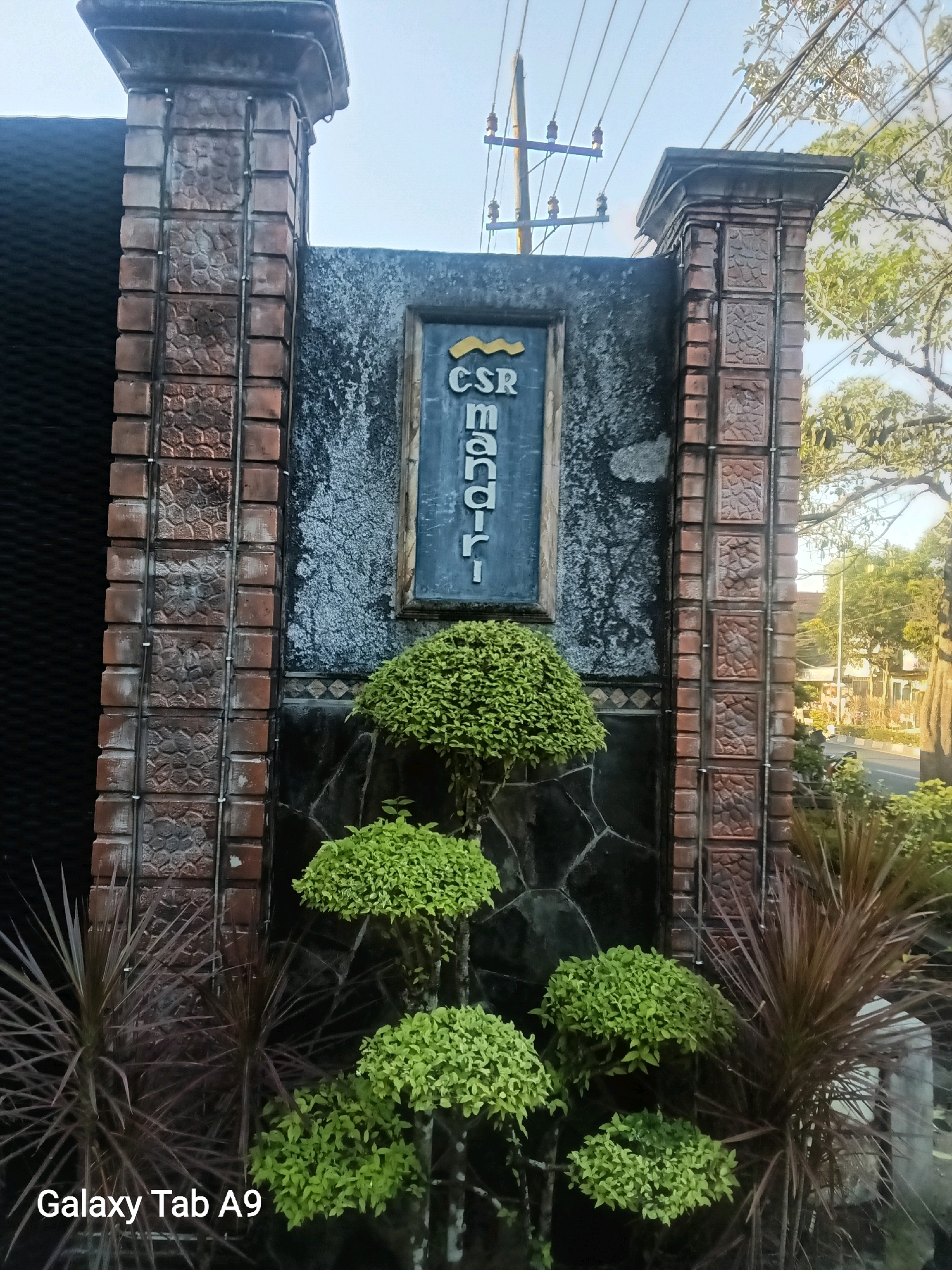 The image displays a sign on a stone wall that reads "CSR Mandiri". The sign is framed by two brick pillars. In front of the sign, there are several potted plants, including some with bushy green foliage and others with spiky, dark red leaves. The background shows a street with trees and buildings. The image is taken from a low angle, looking up at the sign and pillars. The bottom left corner of the image has a watermark that reads "Galaxy Tab A9". Based on the visual cues, the location appears to be the entrance to a building or compound in Kelutan, Indonesia.