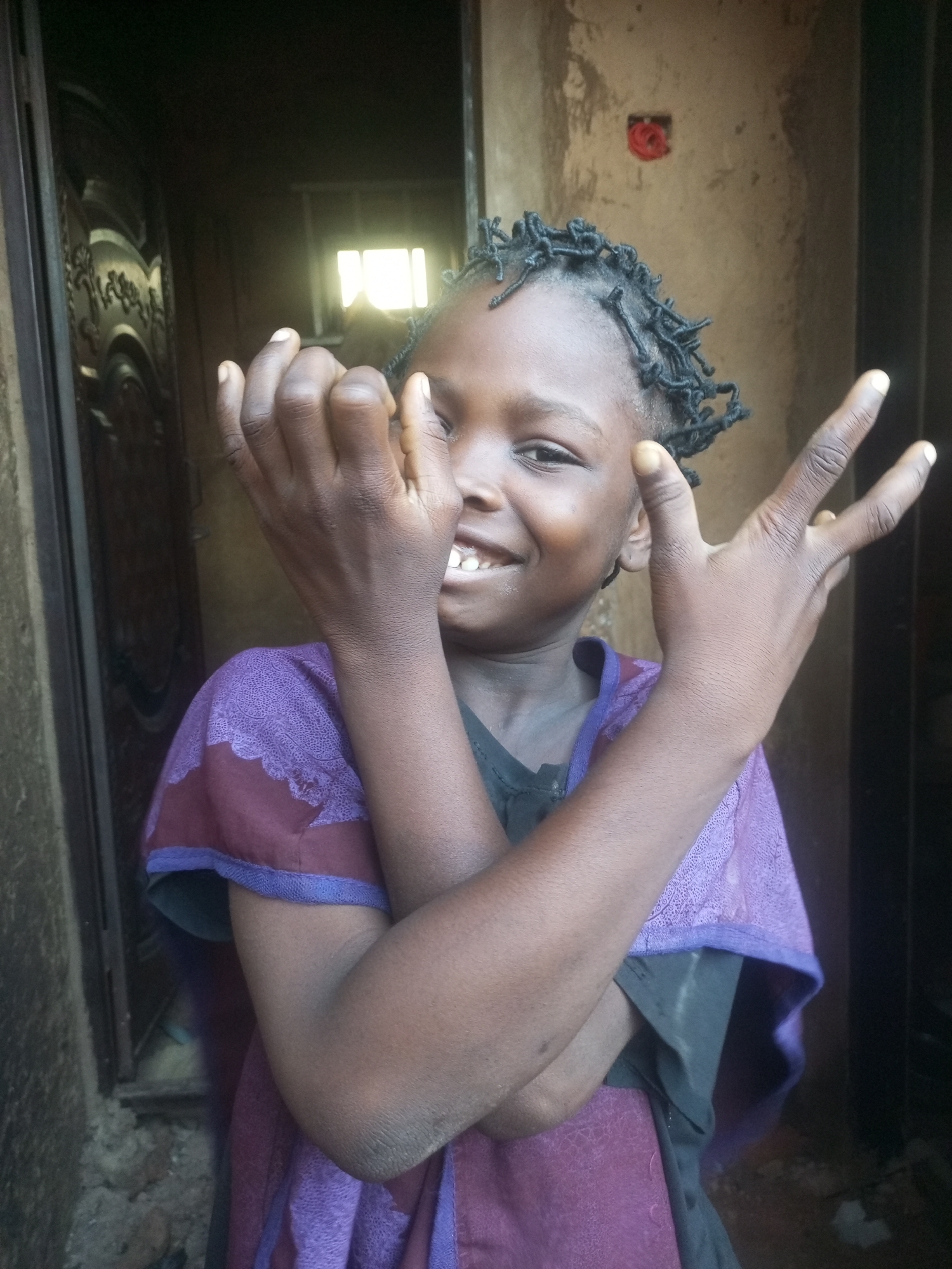 A young child with dark skin and braided hair is standing in front of a rough, textured wall. The child is wearing a purple garment with intricate patterned trim. The child's arms are crossed in front of their chest, and their hands are raised, with fingers extended in a gesture that could be interpreted as a peace sign or a victory pose. The child is smiling, with their teeth visible. Behind the child, a doorway is visible, with sunlight filtering through a window or opening, creating a bright, diffused light. The environment appears to be outdoors, possibly in a rural or less developed area, given the rough walls and simple doorway. The lighting suggests it is daytime. The overall impression is one of a candid portrait of a child in a humble setting. The location is Tempure, Nigeria.