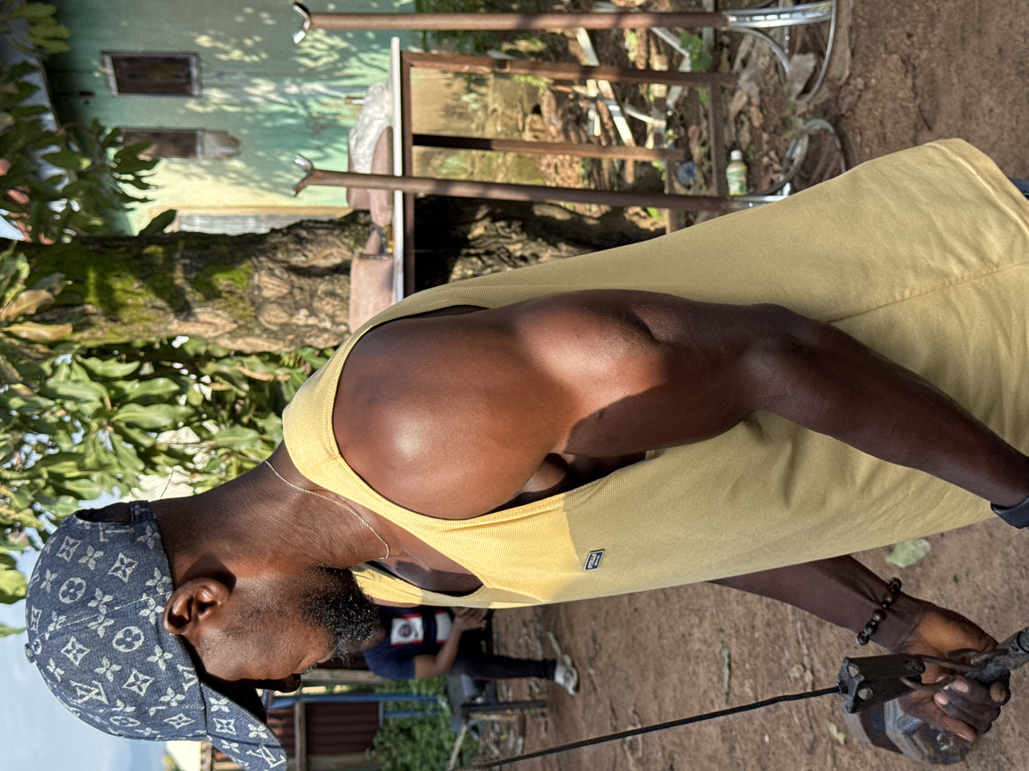 In Akwanga, Nigeria, a dark-skinned man in a denim baseball cap adorned with a Louis Vuitton pattern stands outdoors, facing left in profile. He wears a yellow tank top that reveals muscular arms. He holds a dark object attached to a thin black cord in his left hand, and a beaded bracelet adorns his wrist. In the background, another man in a navy blue t-shirt and black pants sits on a bench, his head bowed. Behind them, a large tree with moss-covered bark stands beside a light green building with two small windows. The ground is covered in dirt and scattered debris, including a plastic bottle and bicycle rims. The scene is illuminated by natural light, casting shadows and highlighting the textures of the environment.