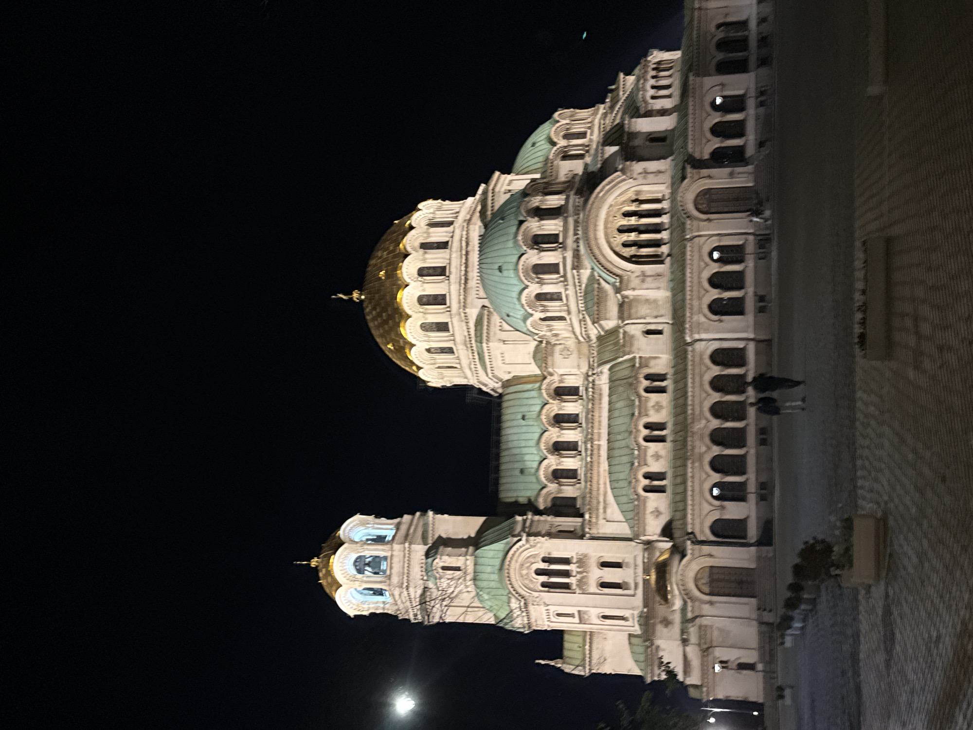 The image shows the Alexander Nevsky Cathedral in Sofia, Bulgaria, at night. The cathedral is illuminated, highlighting its white stone facade, golden domes, and green roofs. The sky is dark. A few people are walking on the paved area near the building. Flower planters line the edge of the pavement. A light source, possibly the moon, is visible in the lower left corner of the frame.