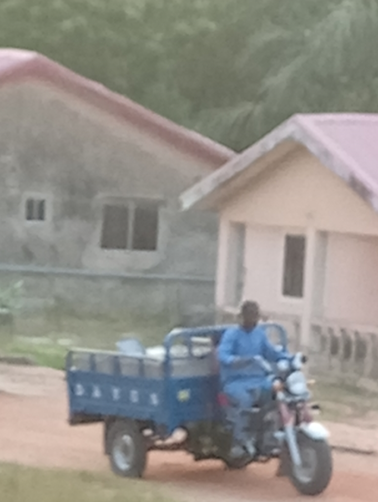 The image depicts a man operating a blue three-wheeled vehicle, possibly a small truck, in front of two buildings. The building on the left is gray with two visible windows, while the building on the right is lighter in color and has a single visible window. The environment appears to be an outdoor, possibly rural, area. The location is Unknown, Unknown.