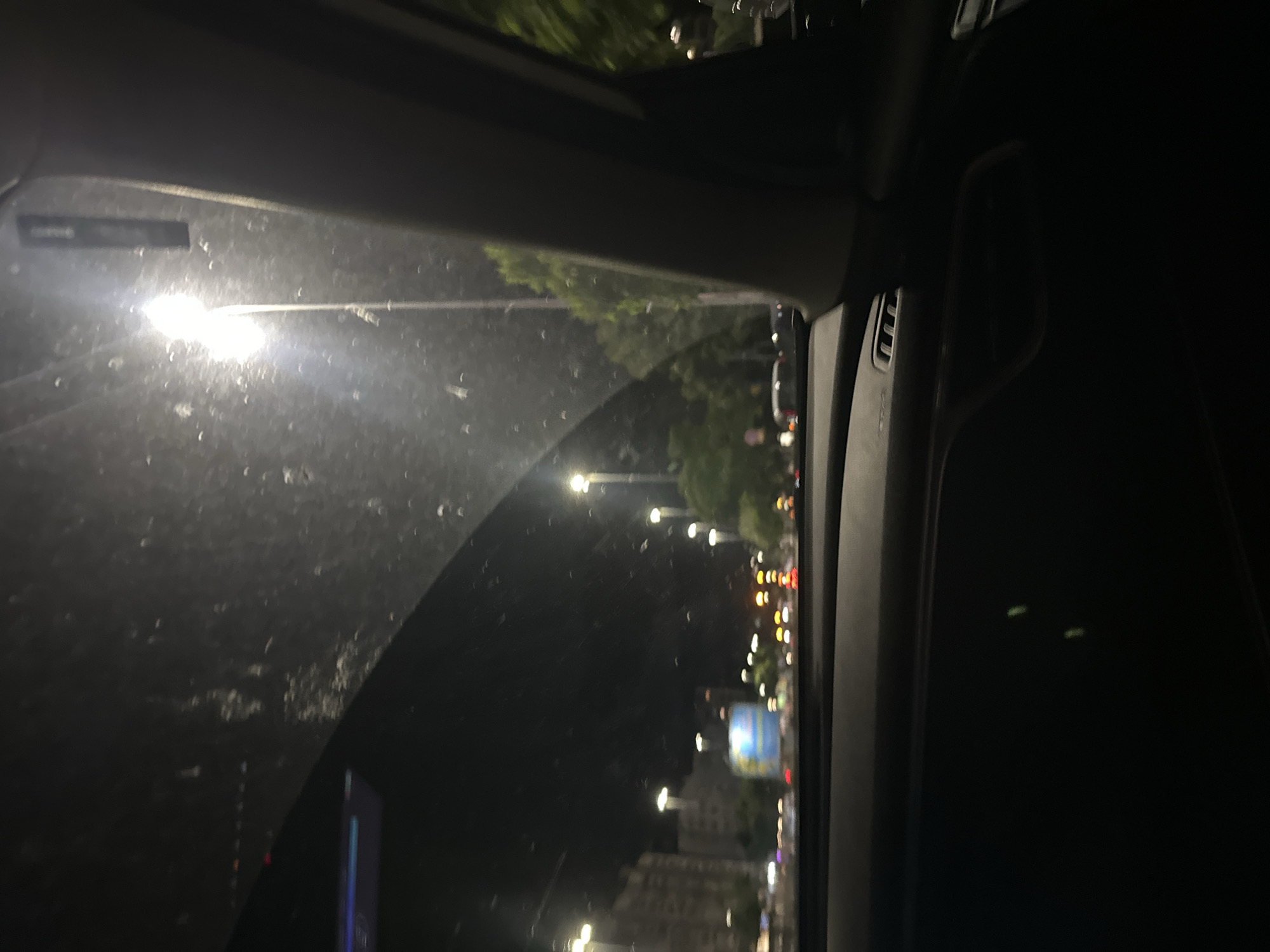 The image shows a view from inside a car at night. The windshield is dirty with water droplets. Outside the car, there are streetlights, buildings, trees, and blurry car lights. The scene suggests a city environment with moderate traffic. The provided crops do not offer specific details about the location in Shumen, Bulgaria, as the image is a general night-time cityscape from a vehicle.