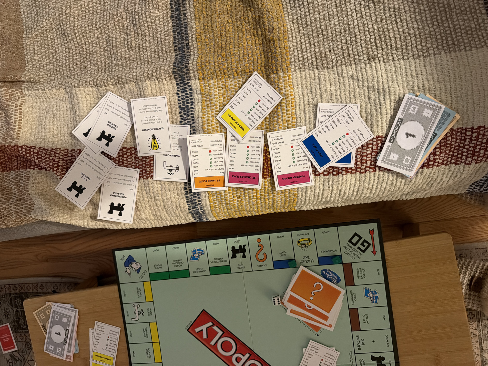 A partially played game of Monopoly is staged on a multi-colored, woven surface, likely a blanket or rug. The Monopoly board, dice, Chance cards, property cards, and Monopoly money are laid out. Property cards, including Atlantic Avenue, Ventnor Avenue, St. James Place, and railroads such as Reading Railroad, are scattered. Monopoly money, in the denomination of one dollar, is visible. The game elements sit atop a wooden surface, probably a table, and the edge of a carpet is visible in the lower right corner. The city of Morton Grove, United States of America, is not evident in the image.