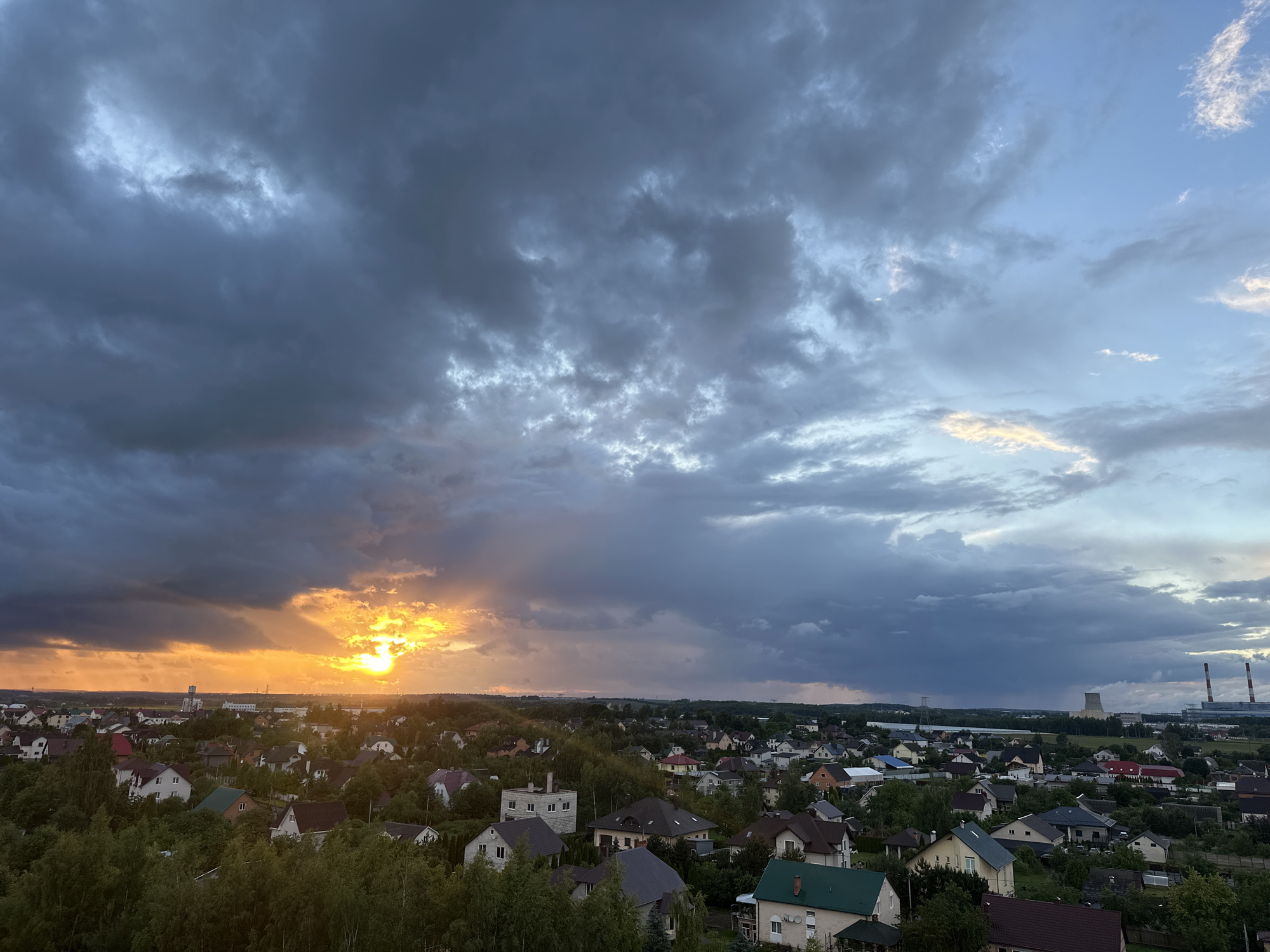 The image shows a panoramic view of a suburban area in the vicinity of Shchomyslitski Sel'ski Sovet, Belarus, during a dramatic sunset. The sky is filled with a mix of dark, heavy clouds and patches of lighter, illuminated sky, creating a stark contrast as the sun sets on the horizon. The sun itself is a bright, intense orb, partially obscured by dark clouds, casting a warm, orange-yellow glow across the landscape. Sunbeams radiate outwards from the sun, cutting through the clouds and illuminating the sky.
Below the sky, a densely populated residential area is visible, characterized by numerous houses with varied roof colors and architectural styles. Many of these houses are surrounded by lush green trees and vegetation, indicating a well-established suburban or semi-rural setting. In the distance, industrial structures, including what appear to be power plant cooling towers and chimneys, are visible on the horizon, suggesting the proximity of industrial facilities to the residential area. The overall impression is one of a vast, developing landscape under a powerful and dynamic sky.
