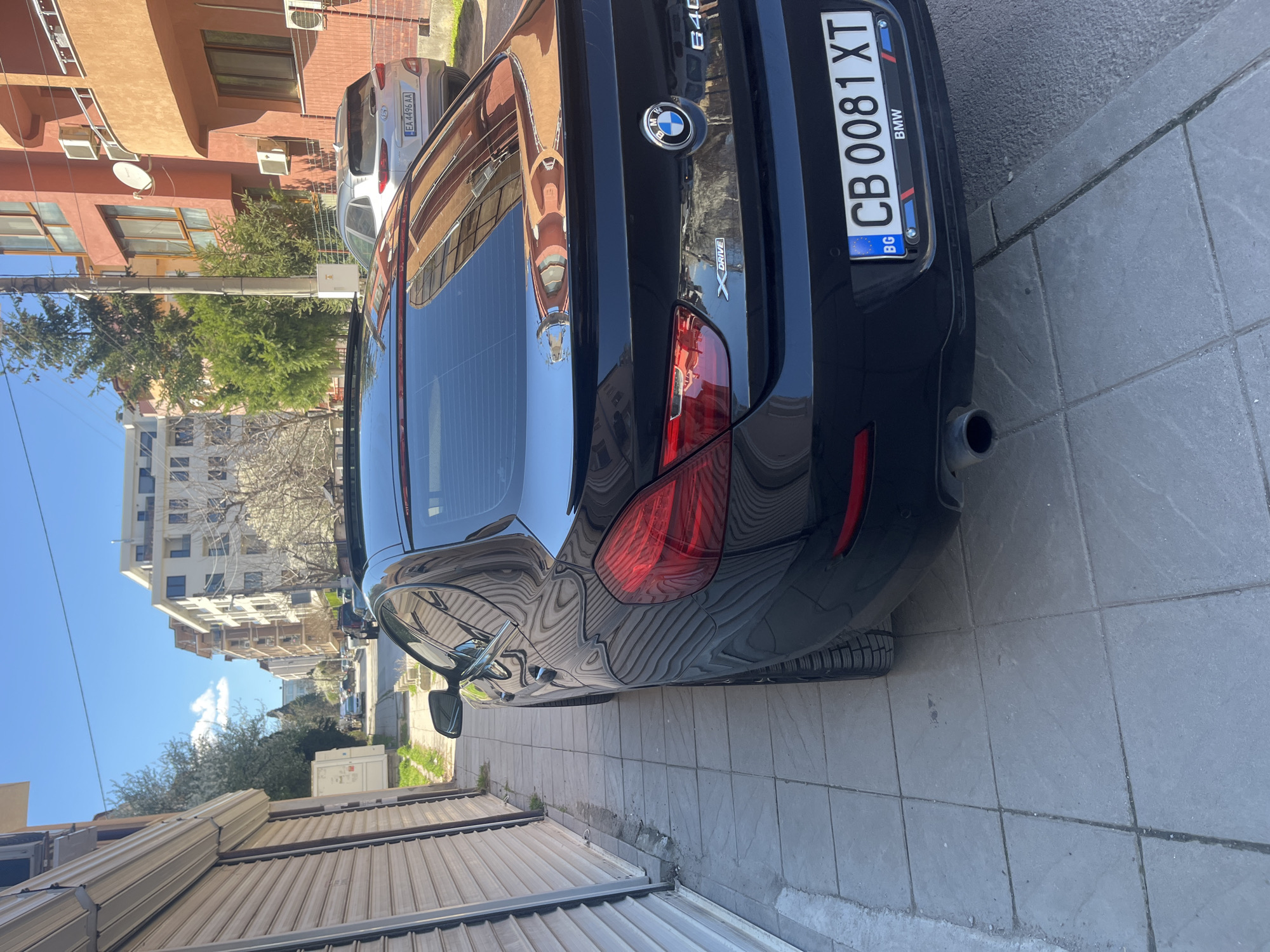 The image depicts the rear of a black BMW 640i xDrive parked on a grey-tiled sidewalk. The license plate reads "CB 0081 XT" and indicates the car is registered in Bulgaria. The BMW emblem is visible on the trunk, along with the "640i" and "xDrive" badges. A silver-colored car is parked behind the BMW. The reflection on the BMW's trunk shows buildings. The surrounding environment consists of buildings, trees, and a blue sky, suggesting an urban setting. The photograph may have been taken in Sofia, Bulgaria, based on the license plate code "CB".