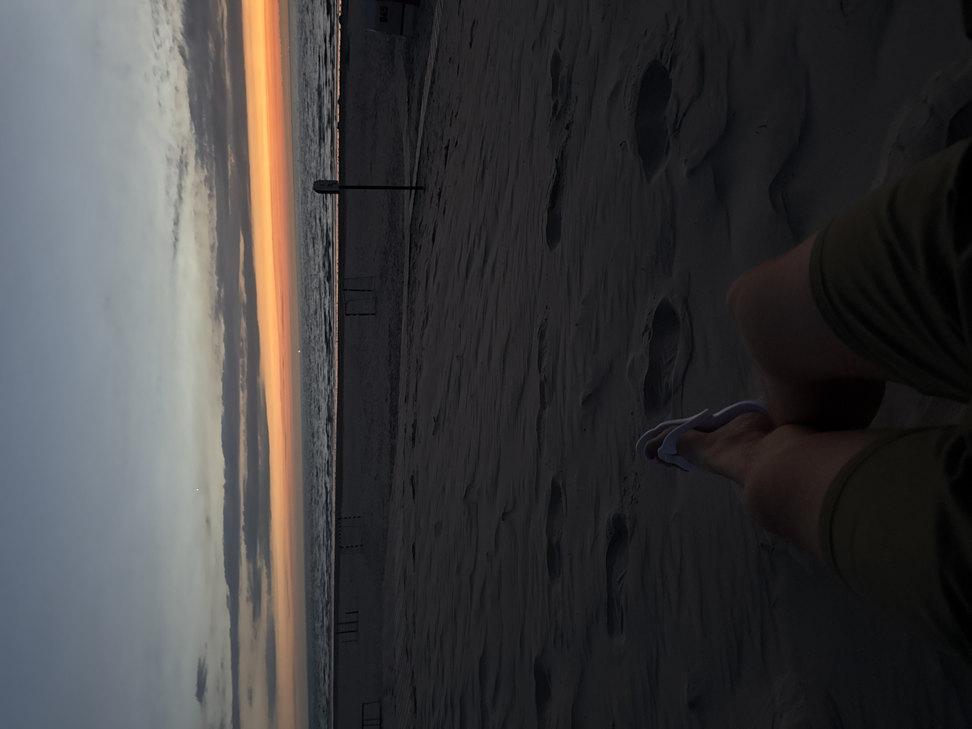 A person's legs and feet are visible in the foreground, with the person wearing green shorts and white flip-flops. They are sitting on a sandy beach in Langeoog, Germany, facing the ocean. The sun is setting, casting an orange glow on the horizon and the clouds. The sky is a gradient of blue and orange. In the background, there are some beach chairs and a soccer net. The overall scene is peaceful and serene, capturing the beauty of a sunset at the beach.