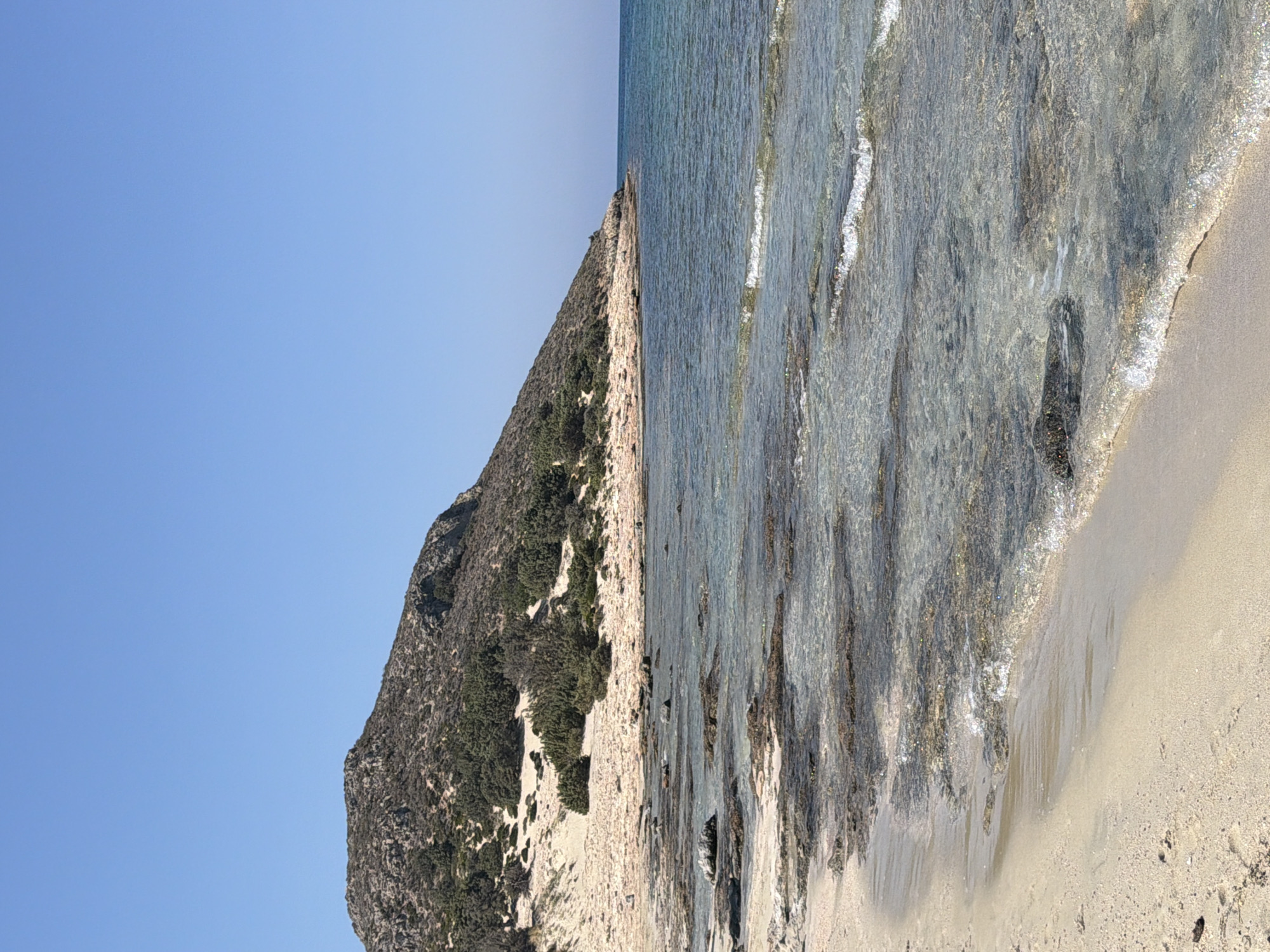 A wide-angle, eye-level shot captures a serene coastal scene under a clear blue sky in an unknown location in Greece. The foreground is dominated by a sandy beach with subtle footprints visible. Gentle waves lap at the shore, their translucent surfaces revealing submerged rocks and seaweed. The water transitions from a pale, sandy bottom near the beach to a deeper, clear turquoise further out. In the midground, a rocky shoreline extends into the sea. The background features a sloping hill covered in sparse, dry vegetation and scattered green shrubs. The hill is predominantly tan and brown, with patches of darker foliage. The horizon is clearly defined by the meeting of the calm sea and the bright blue sky. No people or man-made structures are present in the frame.