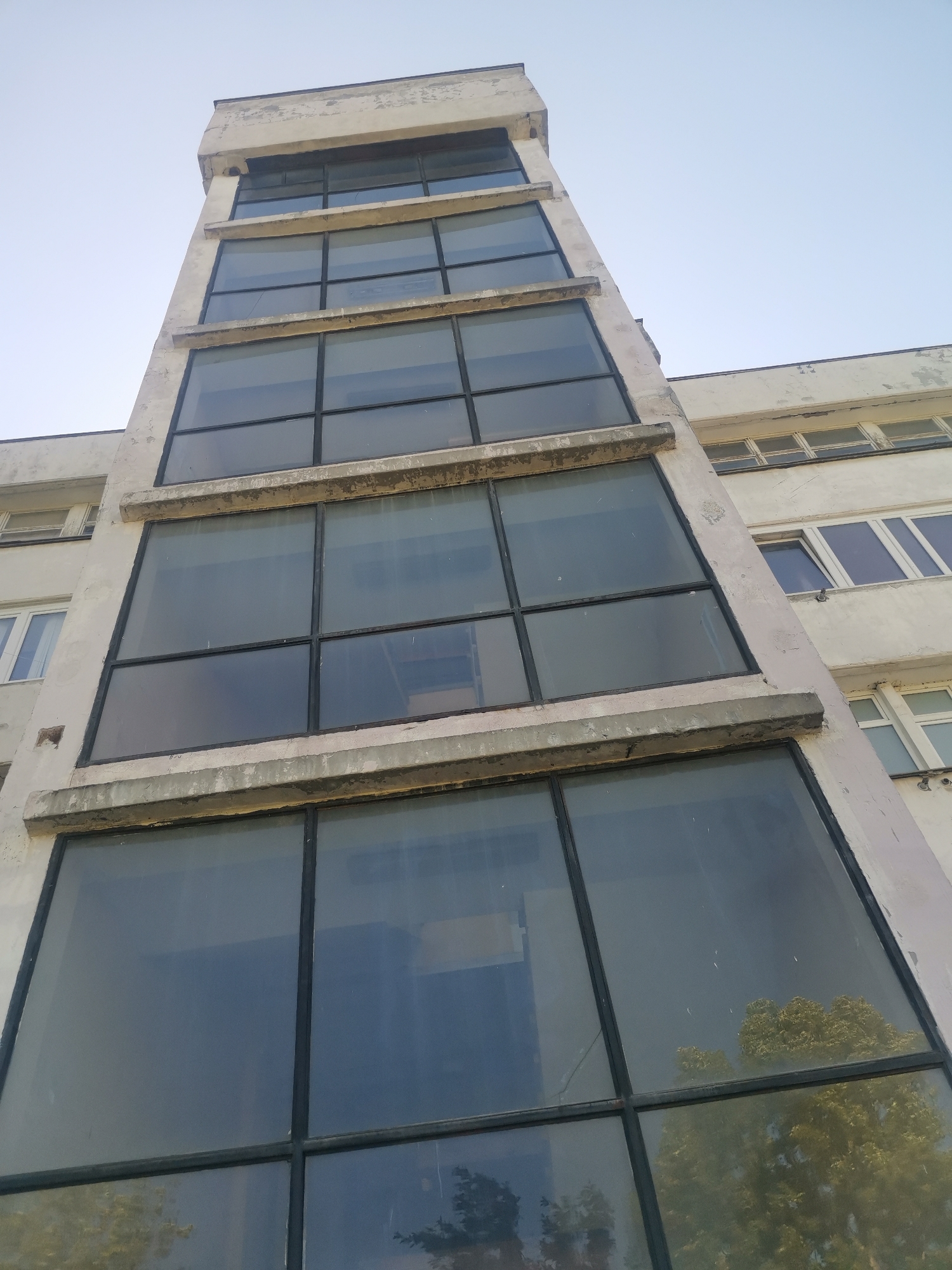 The image shows the exterior of a multi-story building, possibly located in Zhivkovo, Bulgaria. The building features a prominent vertical section, likely an external lift shaft or stairwell, distinguished by a series of large, multi-pane windows framed in a dark metal. This vertical element rises alongside the main structure of the building, which is a lighter color and has smaller, traditional windows. The sky is visible in the background. The building appears to be of a utilitarian, modernist style.