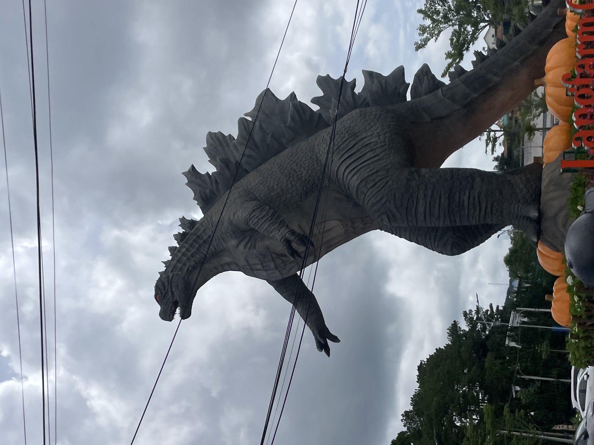 This is an outdoor, eye-level, medium-full shot of a large, gray Godzilla statue positioned in the city of Cam Ly - Đà Lạt, Vietnam. The statue is depicted in a dynamic pose, with its left arm extended forward and its head turned to the left, revealing glowing red eyes. The statue's textured skin and dorsal fins are clearly visible. Several power lines stretch across the frame in front of the statue and the cloudy sky. At the base of the statue, large orange pumpkins are visible, along with some greenery and what appears to be a sign with red lettering that reads "Landfarm". In the background, lush green trees and some buildings are discernible. The overall lighting suggests an overcast day.