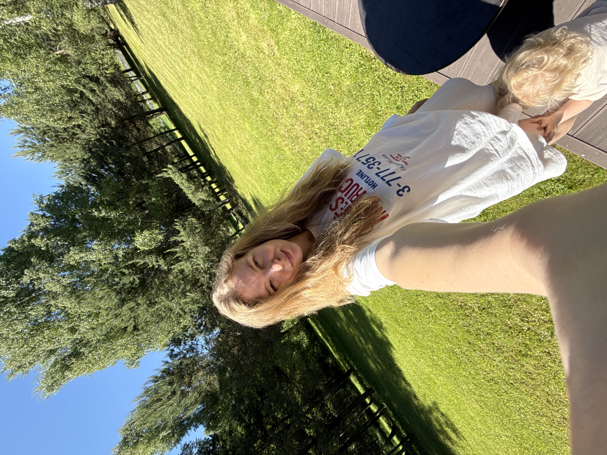The image depicts a bright, sunny outdoor scene in Копнино, Russia. A light-skinned woman with long, light blonde hair captures a wide-angle selfie with her extended right arm, winking her right eye and smiling faintly directly at the camera. She is wearing a loose-fitting white t-shirt bearing the partially visible red and blue text "IES RUCT..." and "HOTLINE 3-777-3571" followed by "Skills Wisdom Your Guide To FUN AT HOME." At the bottom right, a small child with light blonde, curly hair is partially visible, kneeling on the dark wooden planked decking, their hands reaching towards the woman's lower body. A black circular table is also partially visible on the deck to the right. The background features a sprawling, manicured green lawn, brightly lit and stretching into the distance. A dense line of mixed coniferous and deciduous trees forms the backdrop, casting long shadows across parts of the grass. The sky in the upper left is clear and bright blue.